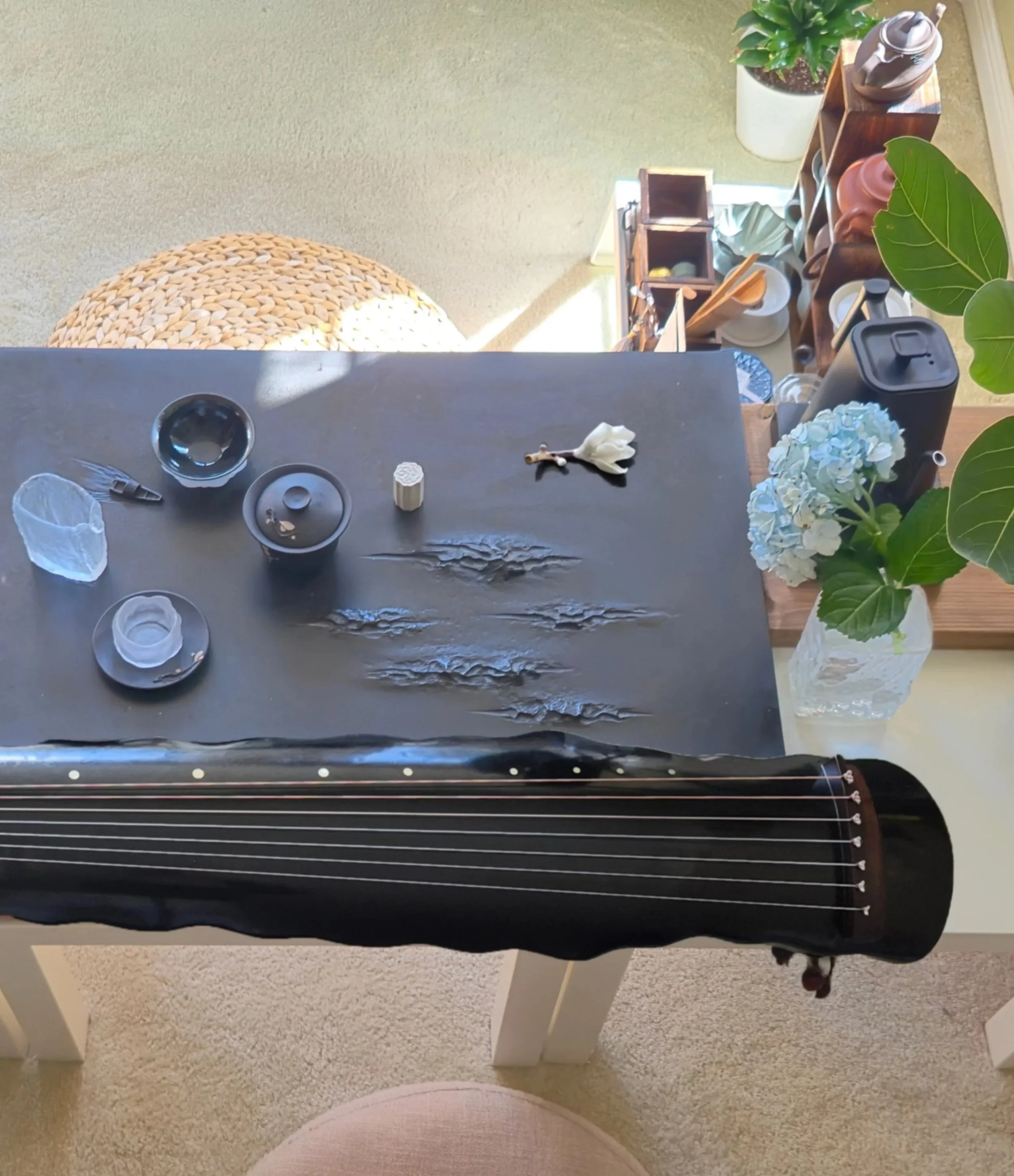 Top-down view of a black tabletop with various objects including a black guitar headstock, a white flower in a vase, small bowls, a plant, a black container, and a wooden organizer, with a wicker placemat and beige carpet visible nearby.