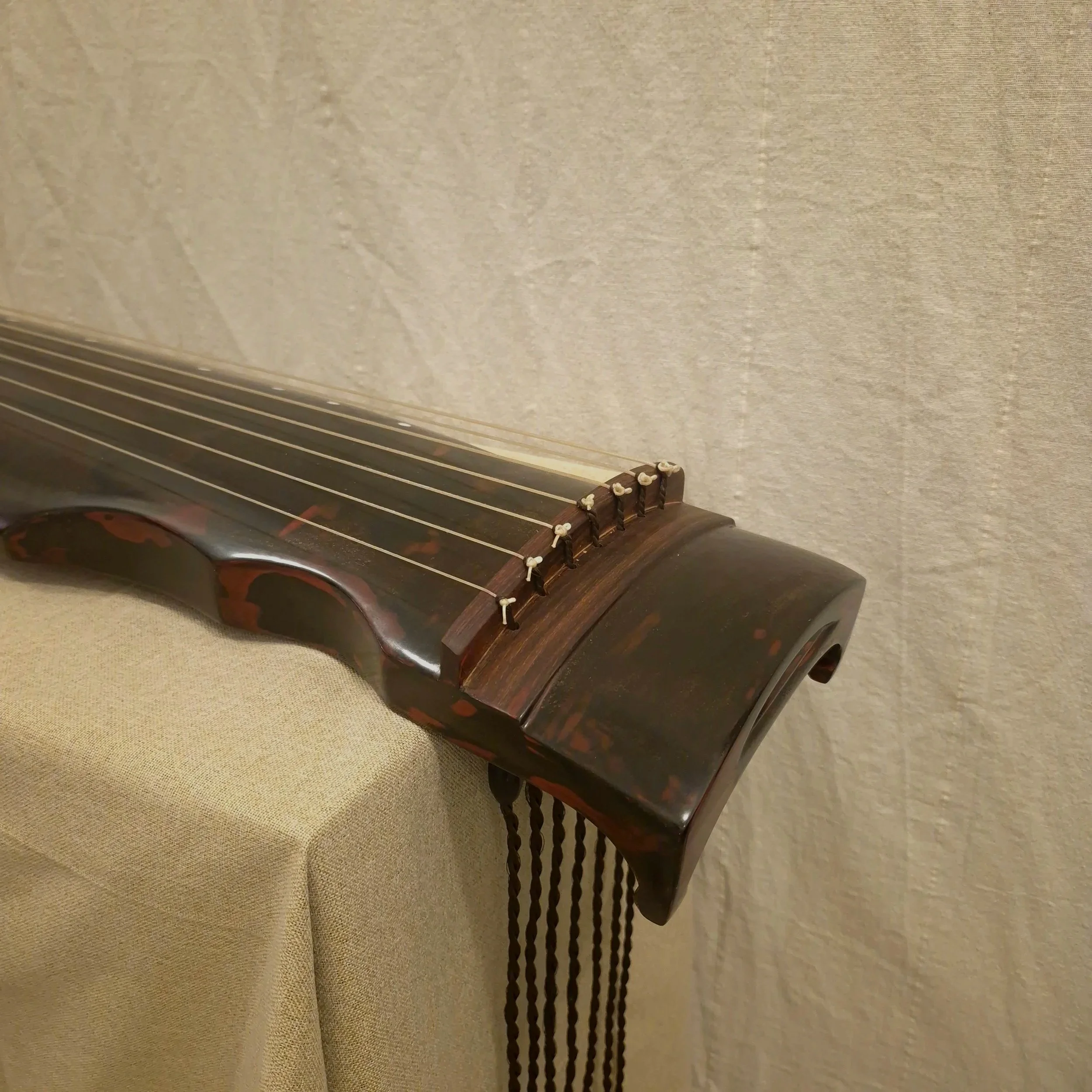 Close-up of the neck and fretboard of qin, showing tuning pegs and strings, resting against a beige textured surface.