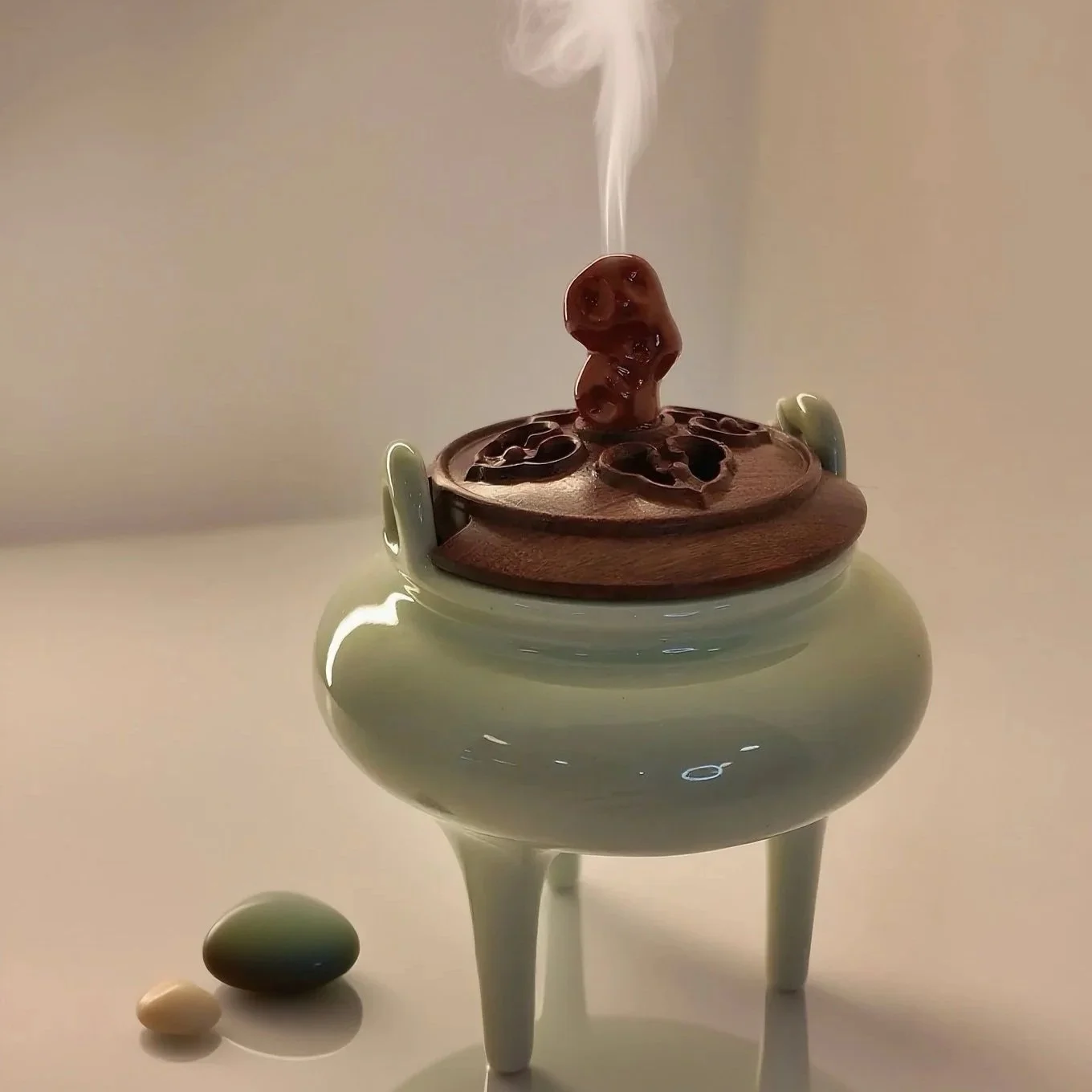 A steaming ceramic aroma diffuser with three legs, containing dark wooden aroma chips on top, and a single block of herbal incense rising smoke. Two stones, one white and one dark green, are placed beside it on a white surface.