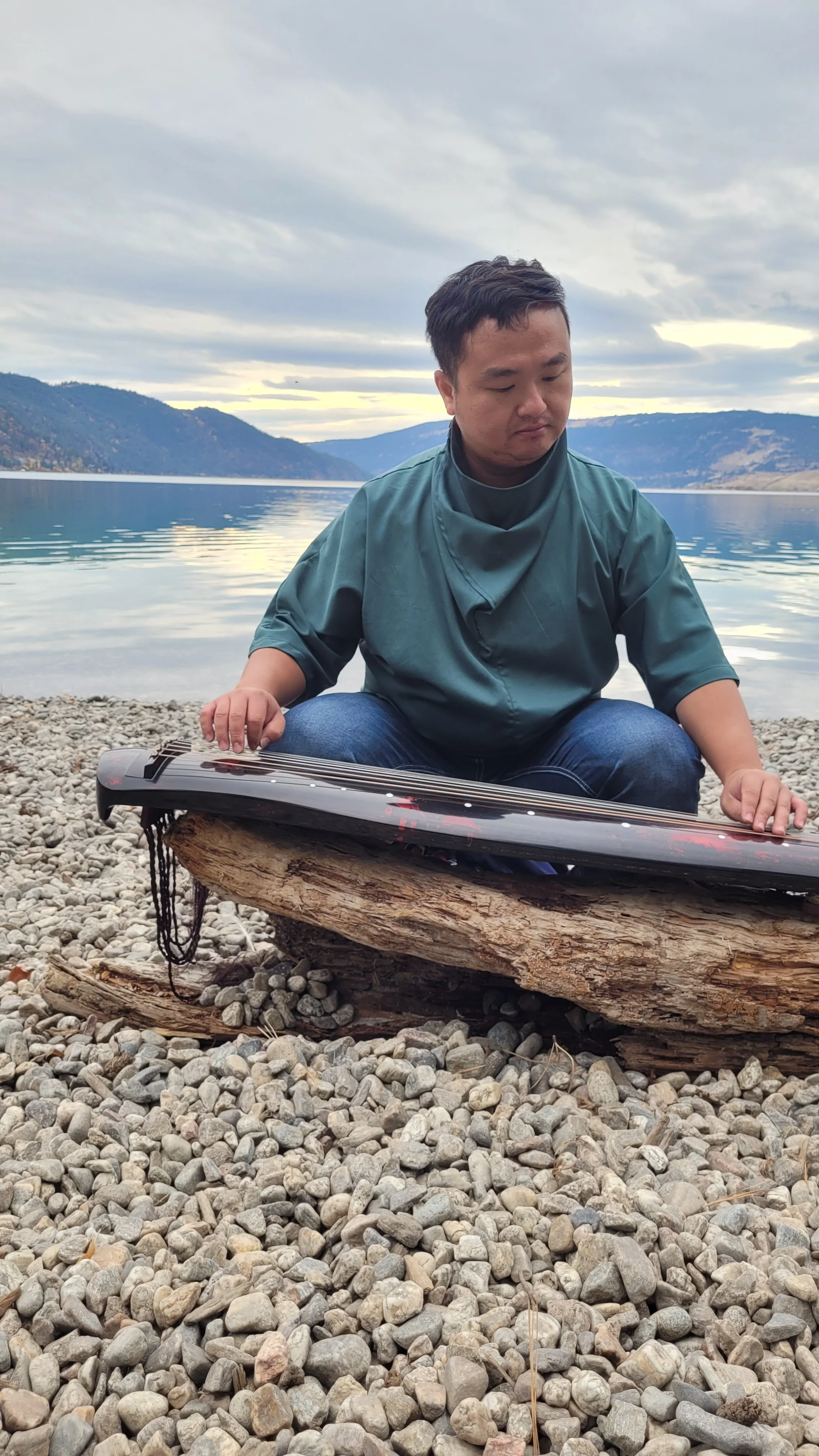 Atom Wang in a teal shirt sitting on a log at  Kalamalka lake, with mountains and cloudy sky in the background, holding a black paddleboard or kayak.