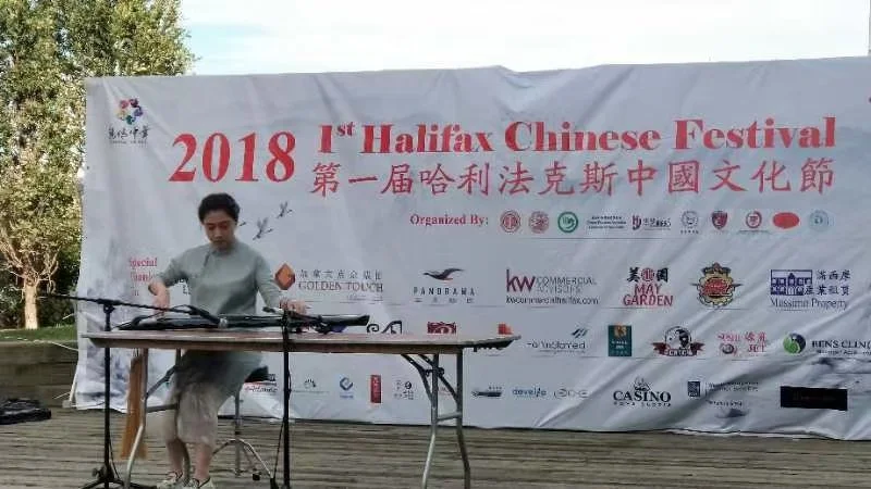 A performer playing a traditional Chinese musical instrument at the 2018 Halifax Chinese Festival, with a large banner featuring event details and sponsor logos in the background.