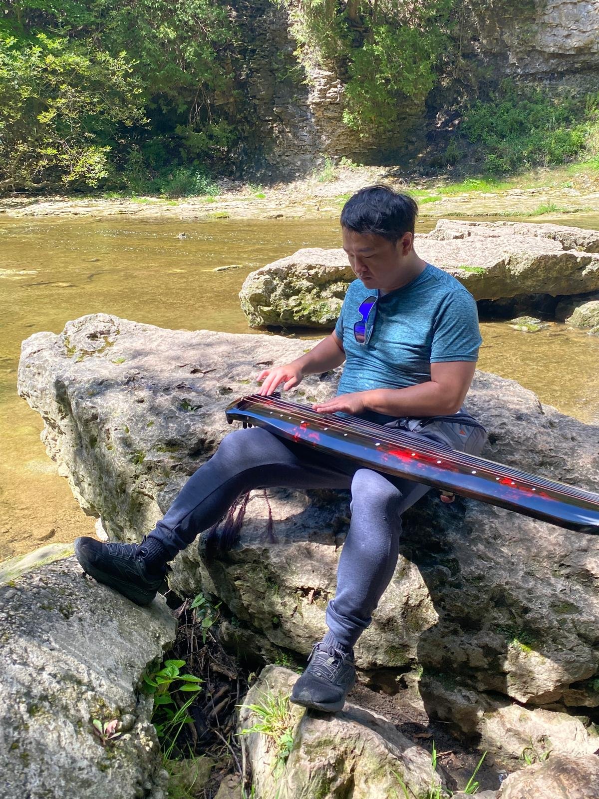 Atom Wang sitting on a large rock by a shallow stream, playing qin on his lap surrounded by green trees and rocks.