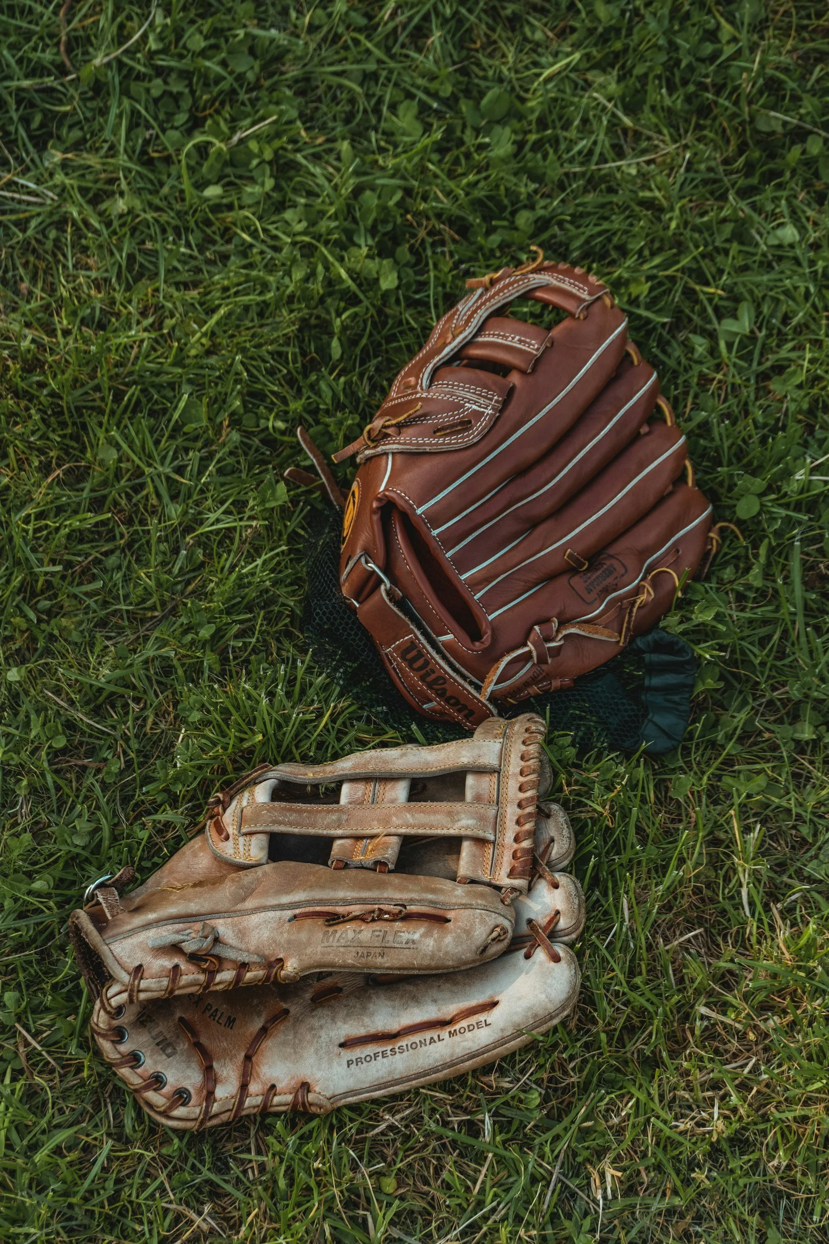 A worn baseball glove and a new baseball glove lying on green grass.