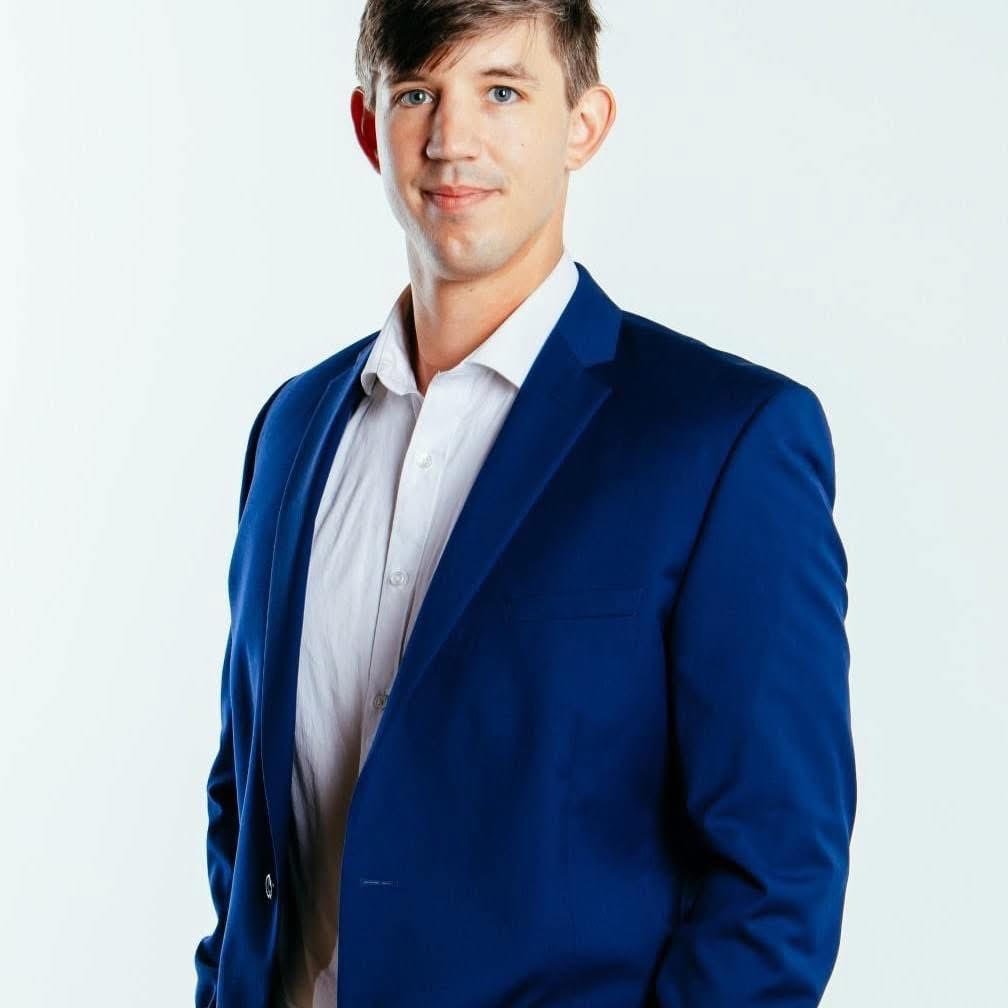 A young man with short brown hair, dressed in a white button-up shirt and a blue blazer, standing against a plain white background.
