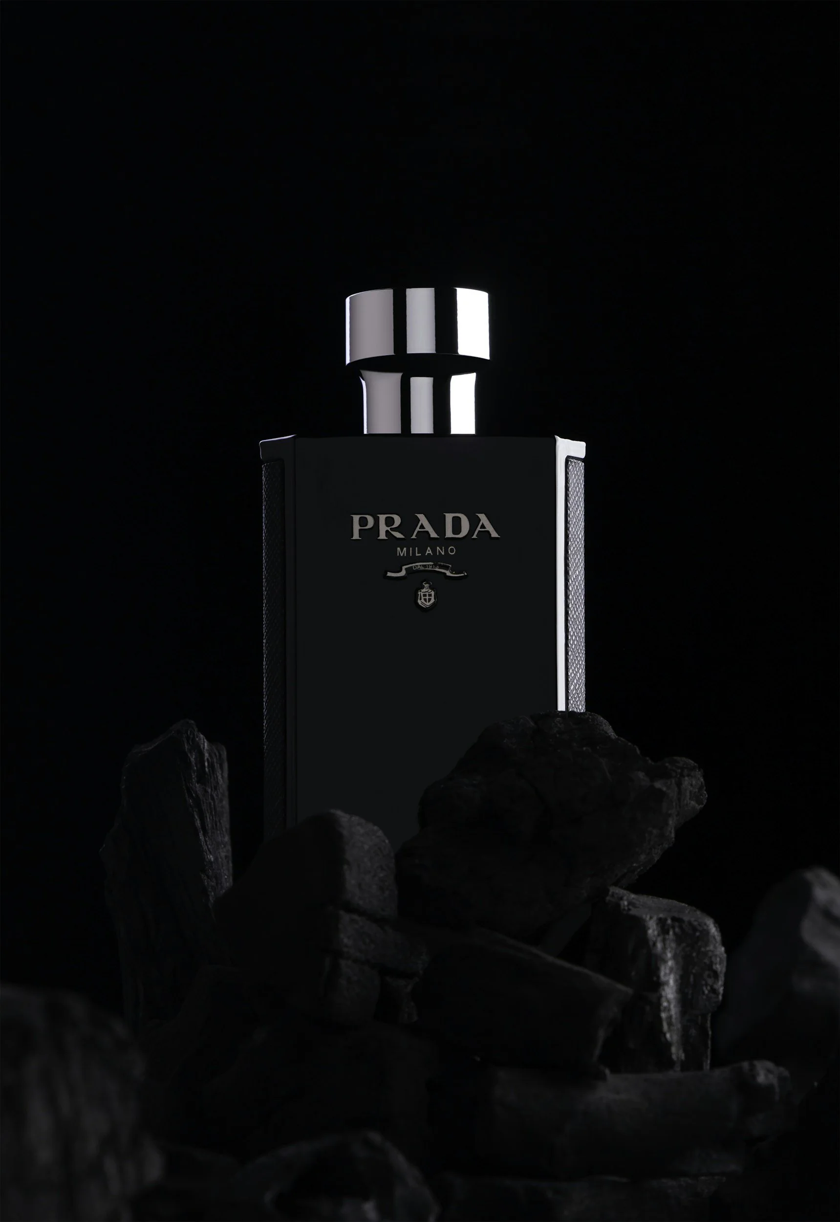 A black Prada Milano perfume bottle placed among dark rocks against a black background.