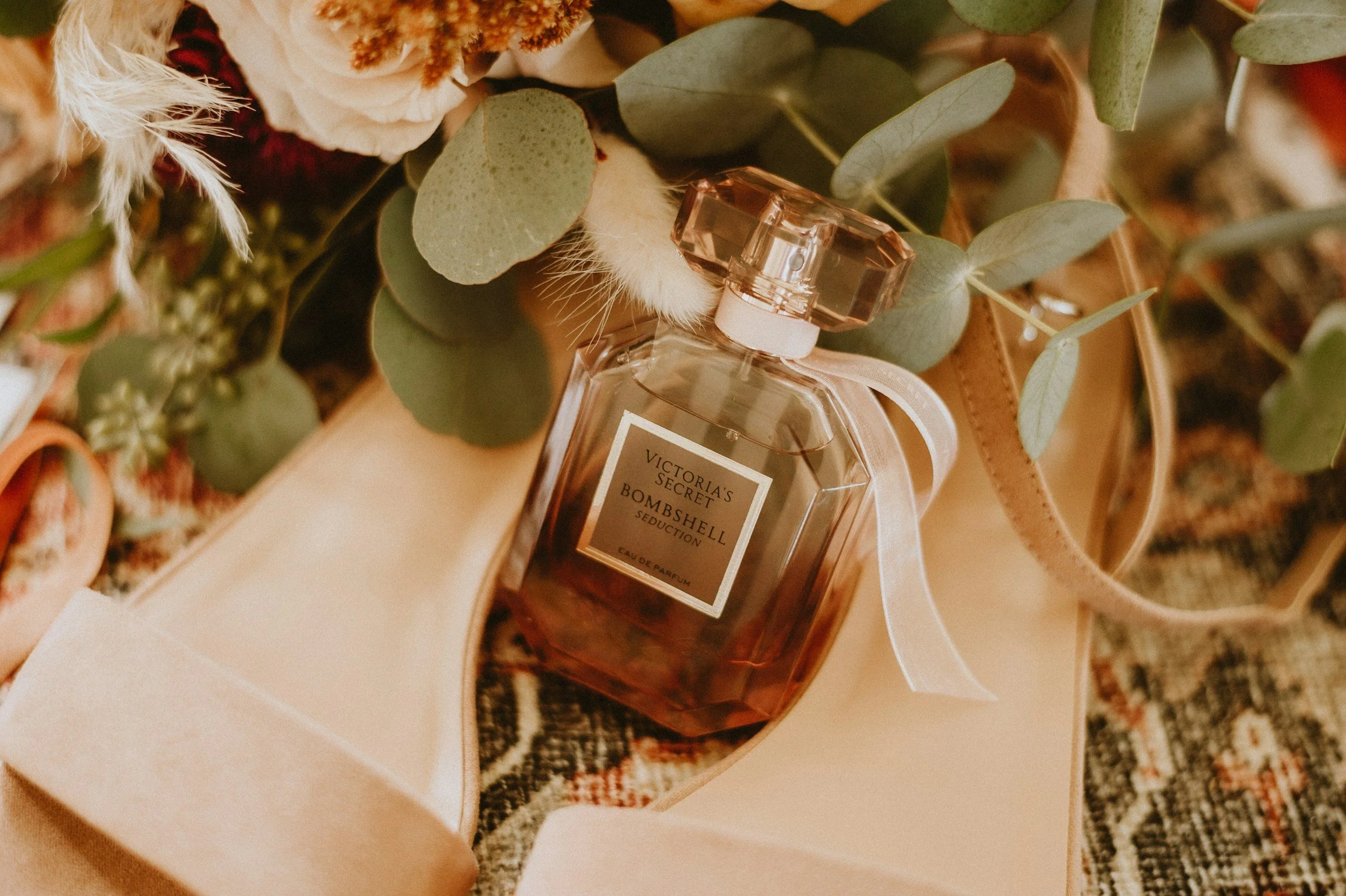 A Victoria's Secret Bombshell Seduction perfume bottle surrounded by flowers, greenery, and ribbons on a decorative fabric surface.