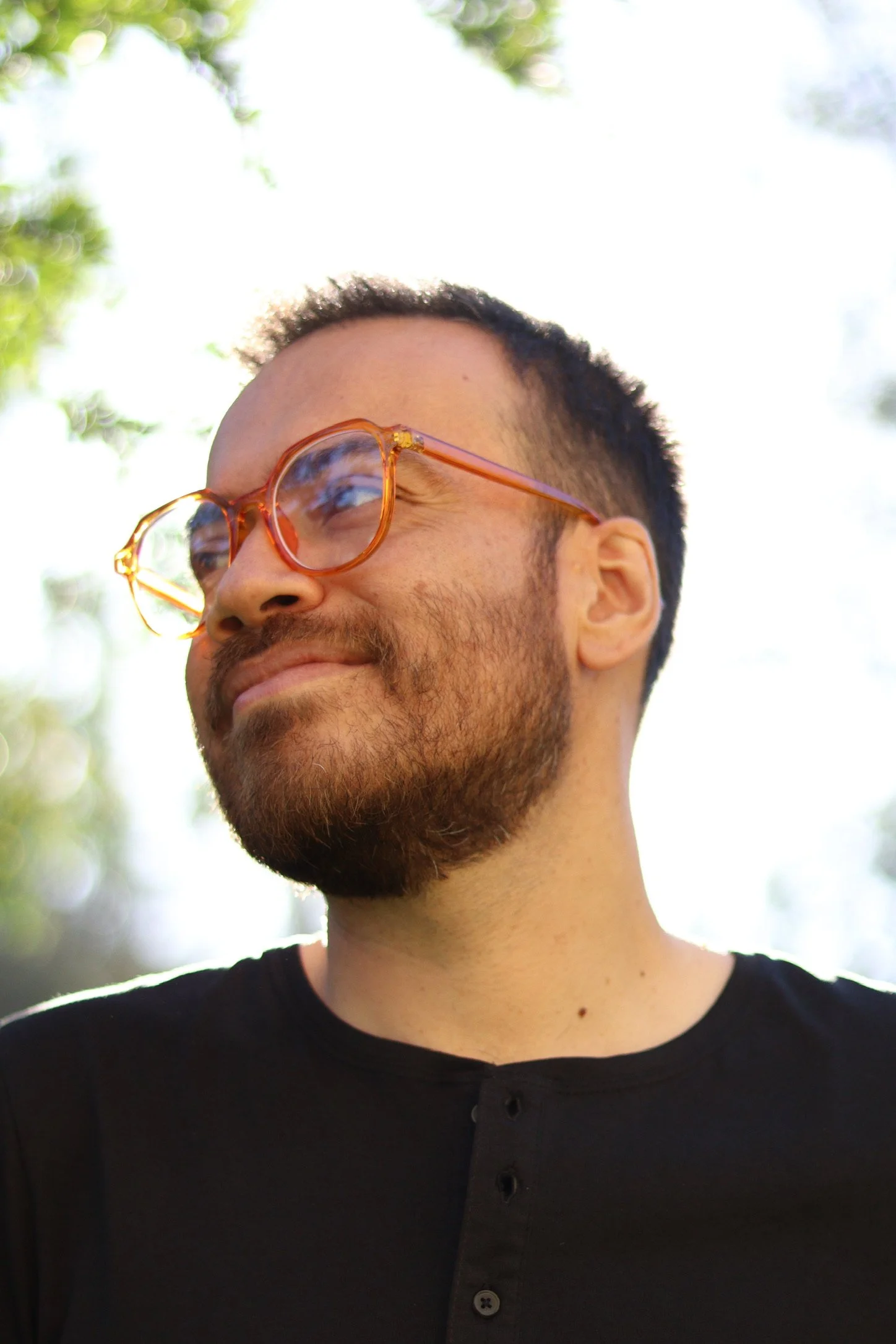 Hombre con barba y gafas naranjas, sonriendo y mirando hacia la izquierda, con fondo de árboles y cielo.