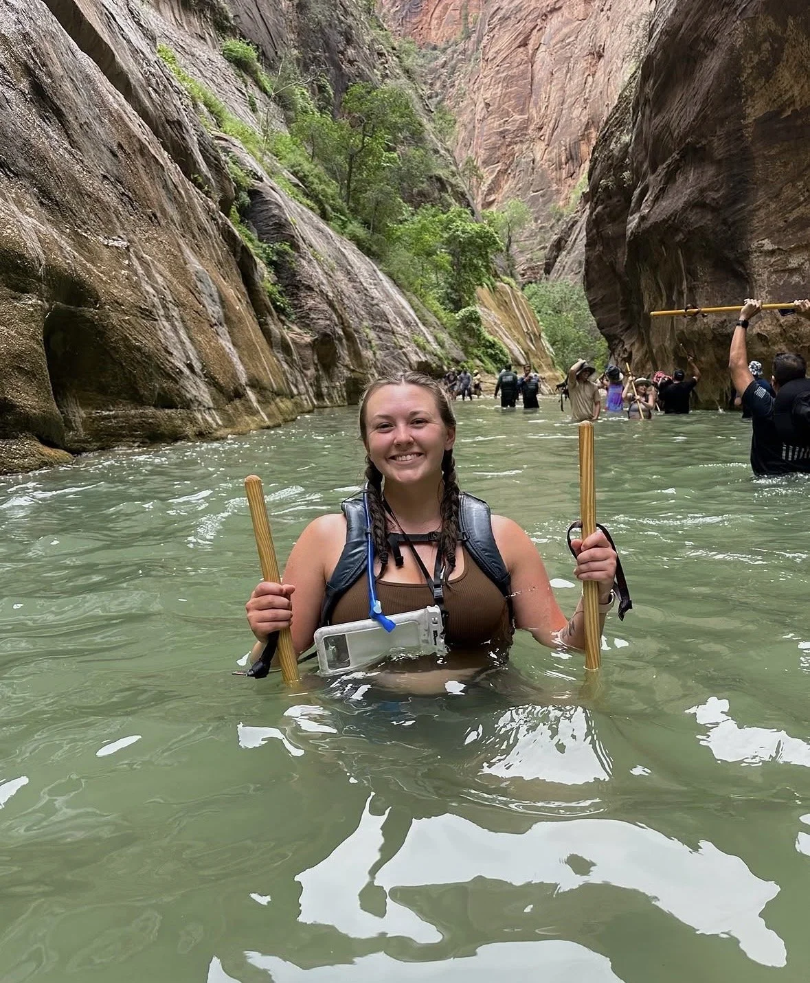 A smiling woman in a swimsuit and life jacket wading in a river inside a canyon, holding a wooden stick in each hand. Other people are visible in the background, also wading through the water between tall canyon walls with greenery.