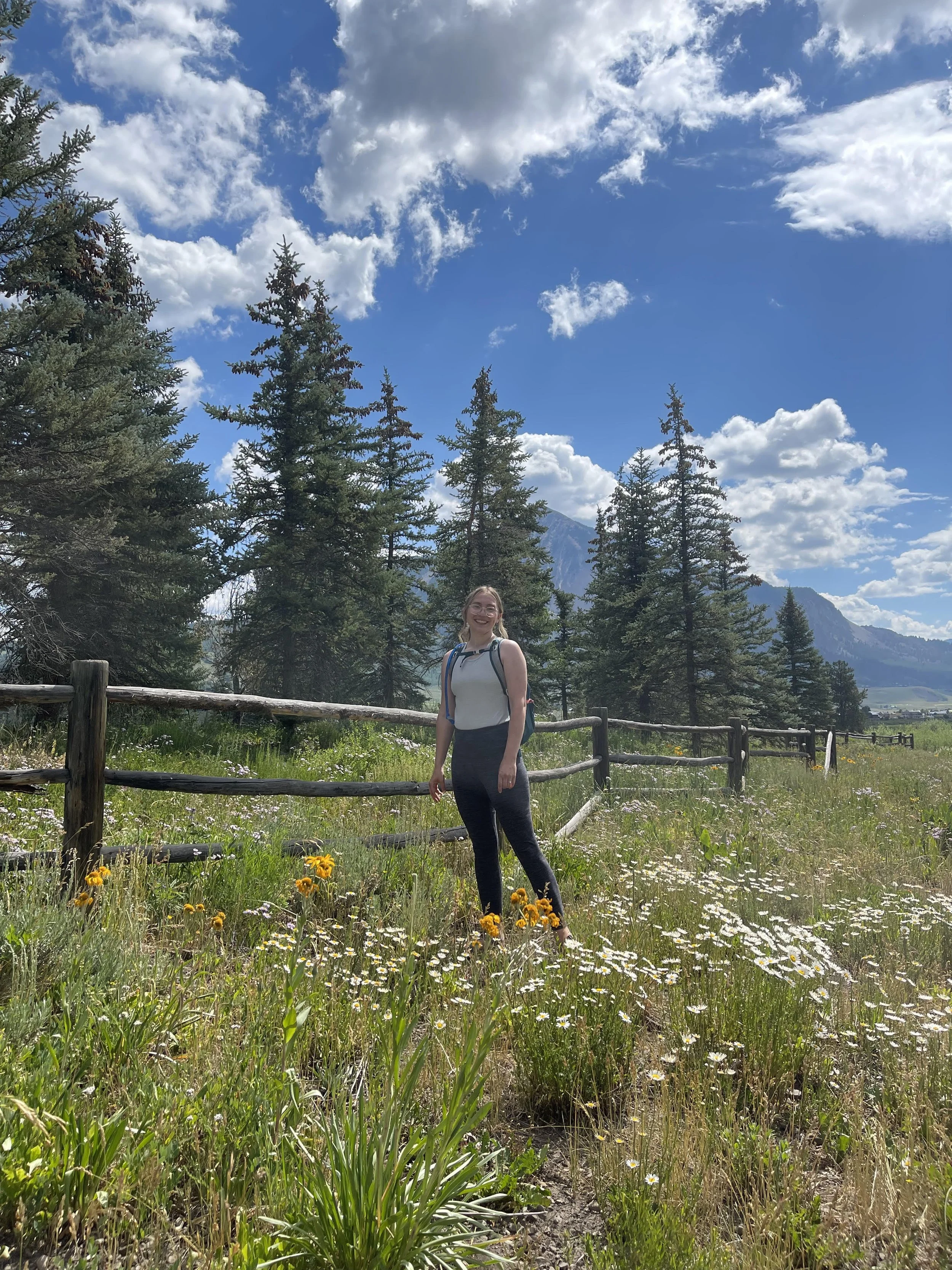 A woman outdoors in a grassy field with wildflowers and tall pine trees, mountains in the background, blue sky with scattered clouds.