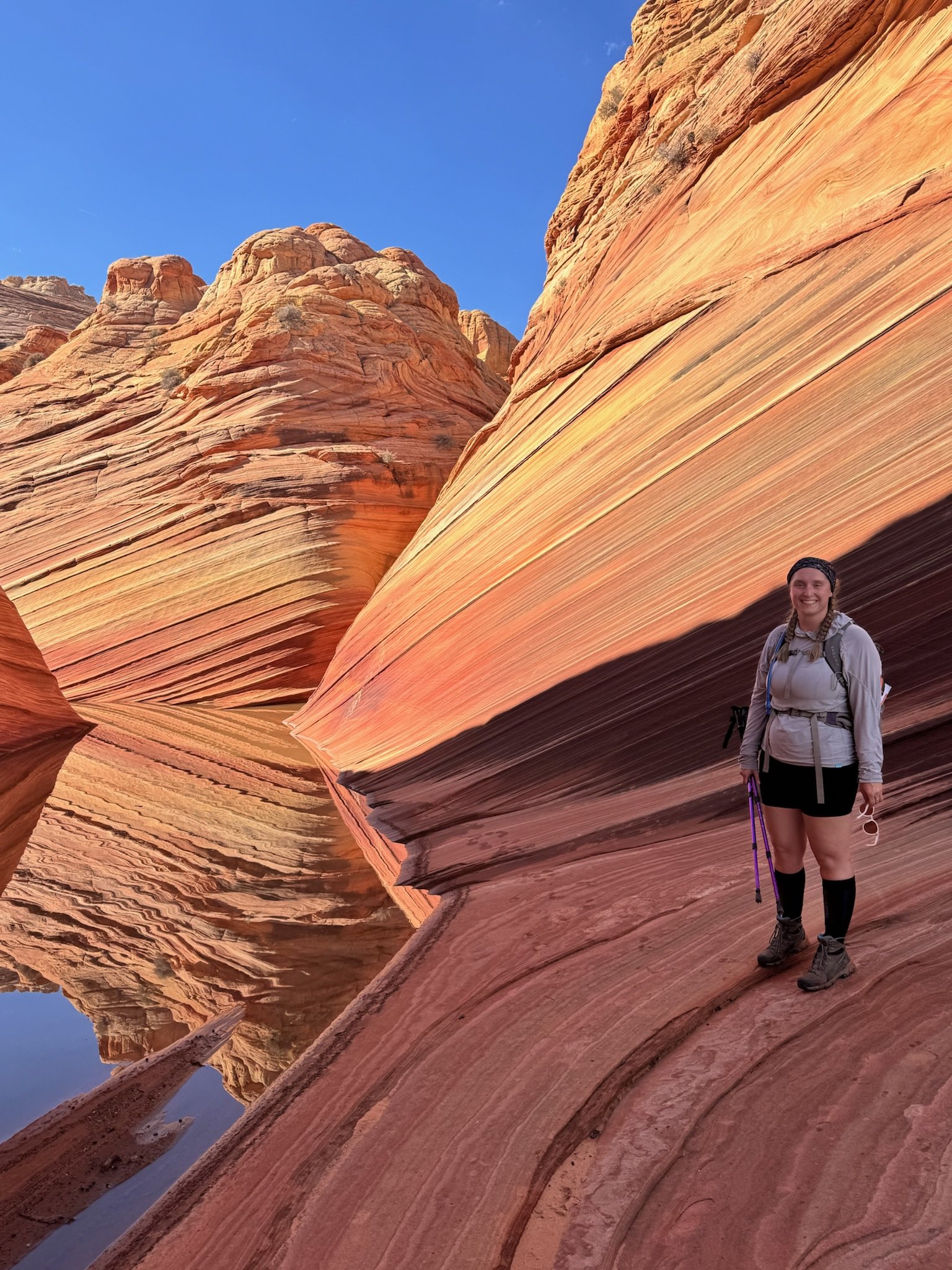 A woman standing inside a slot canyon with colorful, layered sandstone walls and a small pool of water at her feet, wearing outdoor hiking gear and smiling.