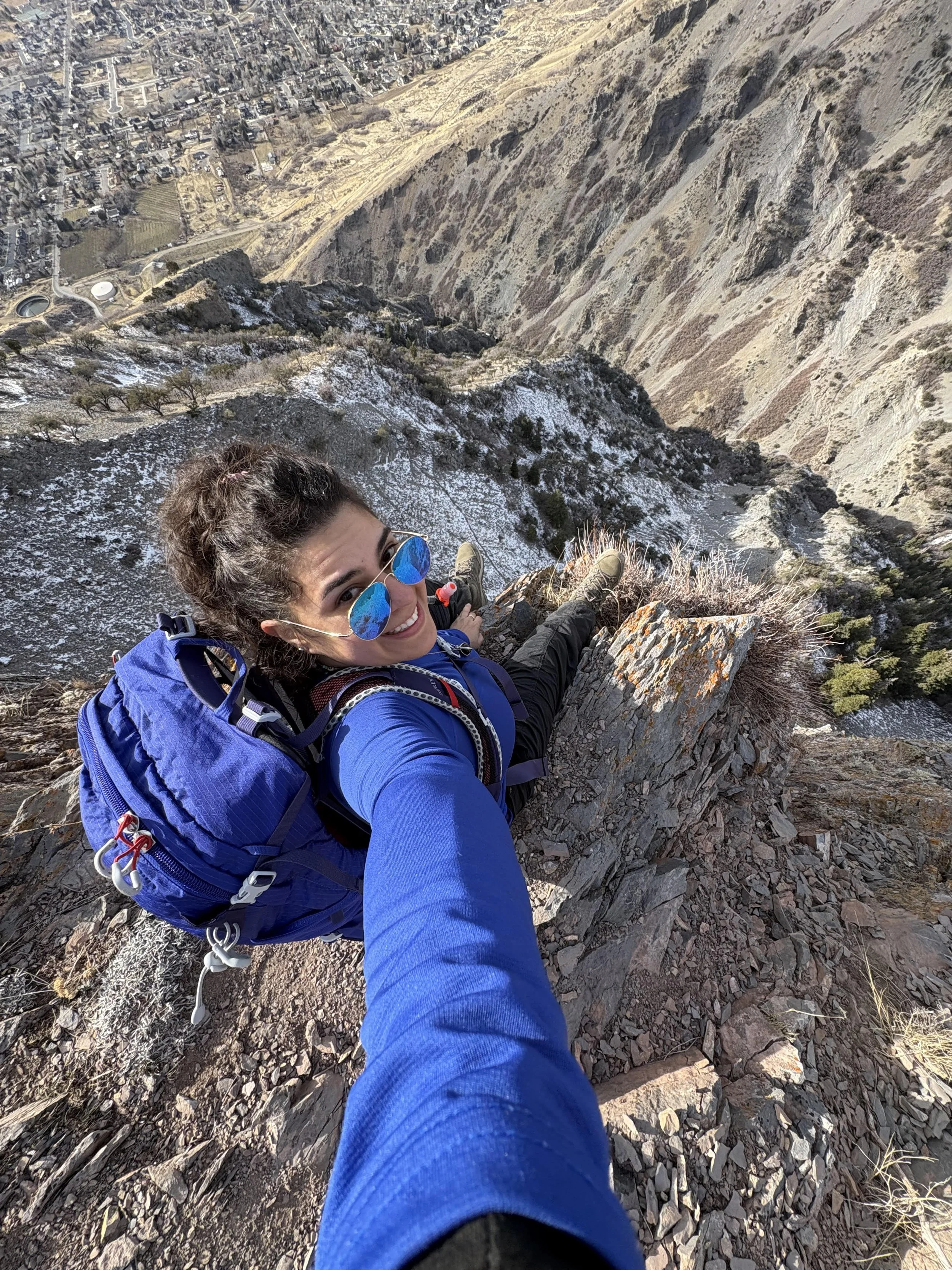 A woman with sunglasses taking a selfie from a mountain ledge with a city below and a steep rocky slope in the foreground.