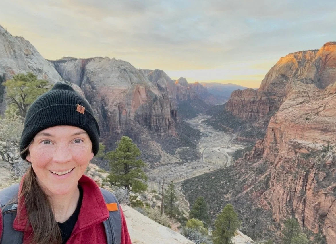 A smiling woman standing on a hillside overlooking a canyon with cliffs, trees, and a river in the distance at sunset.