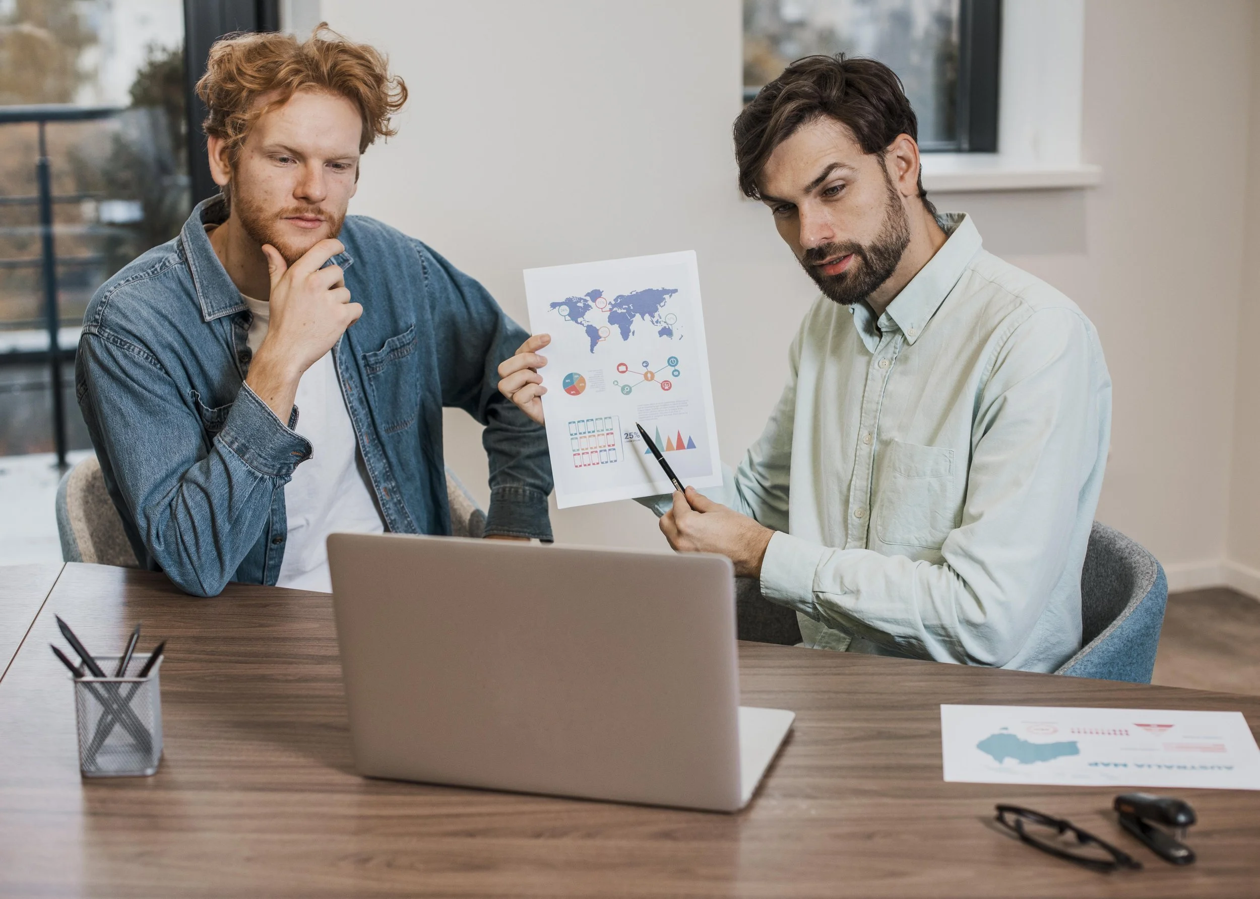 Two men working together in an office, looking at a printed infographic with a world map, charts, and graphs during a meeting.