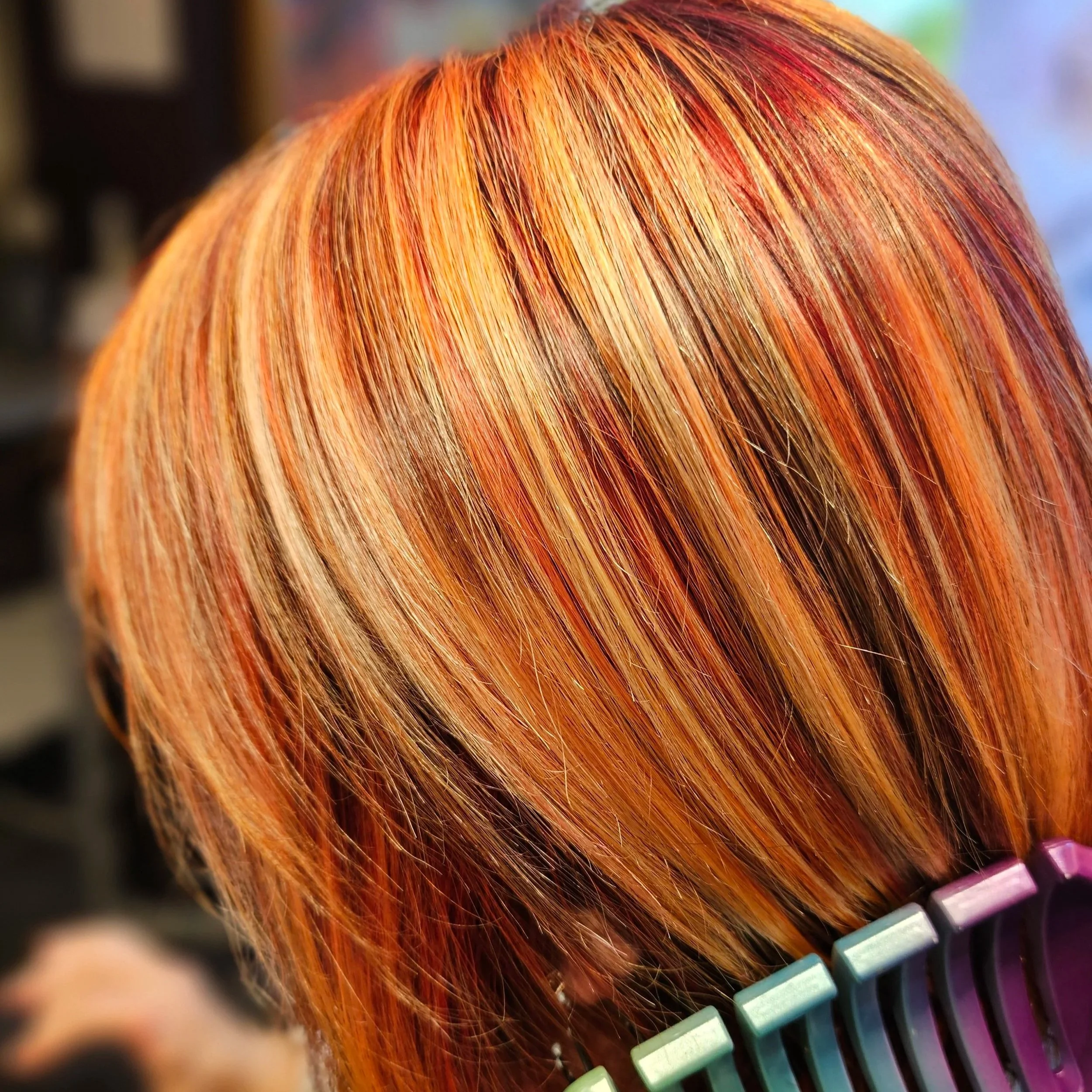 Close-up of a person's hair dyed in vibrant shades of orange, red, and blonde, styled in a straight bob cut, with a hairbrush at the bottom right of the image.