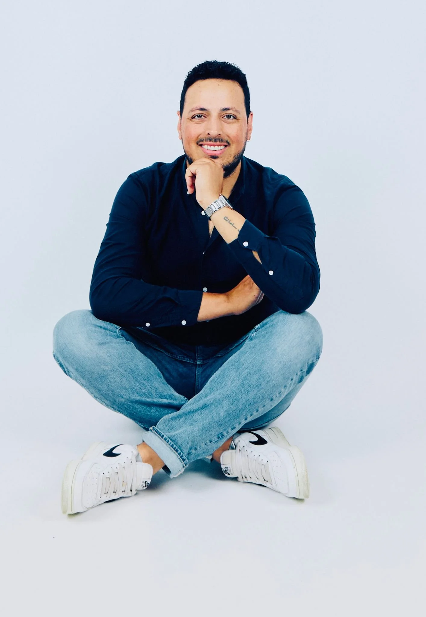 Man sitting cross-legged on the floor, smiling, wearing a dark blue shirt, light blue jeans, and white sneakers, with a white background.