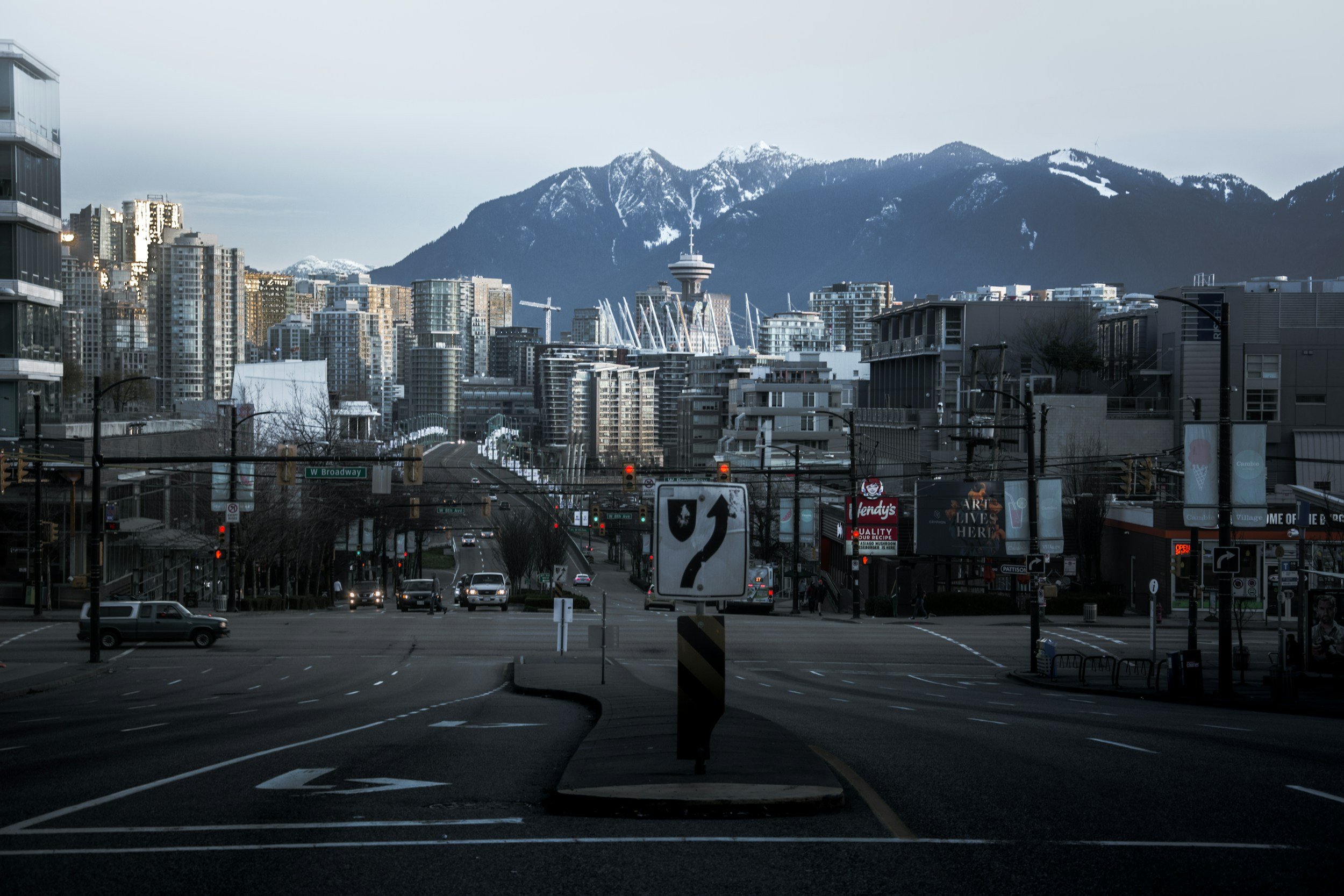 Empty city street intersection with tall buildings, mountains in the background, and a cloudy sky.