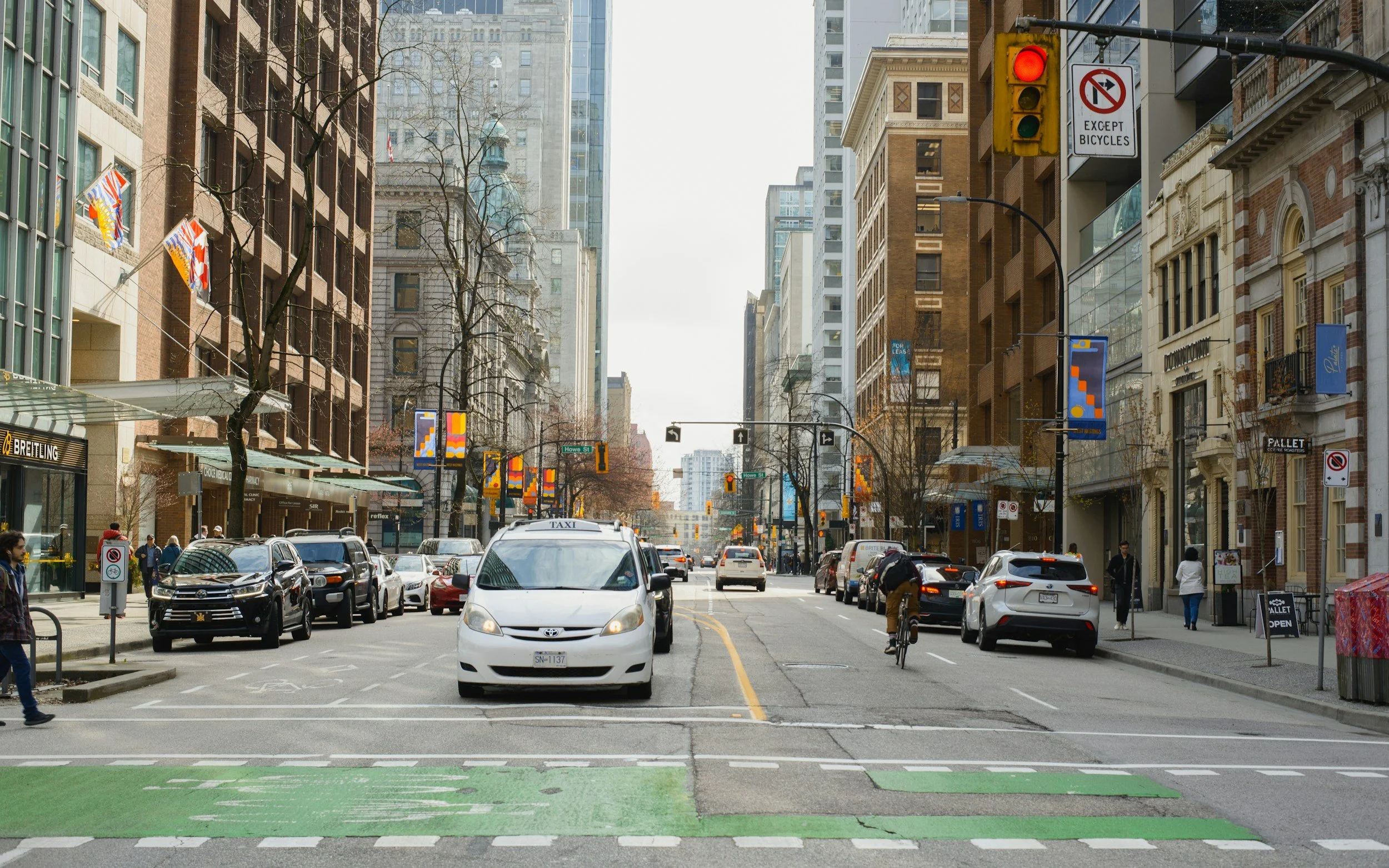 Downtown city street showing cars, taxis, and a cyclist with tall buildings and storefronts on both sides, some pedestrians walking on sidewalks, with traffic signals and street signs.