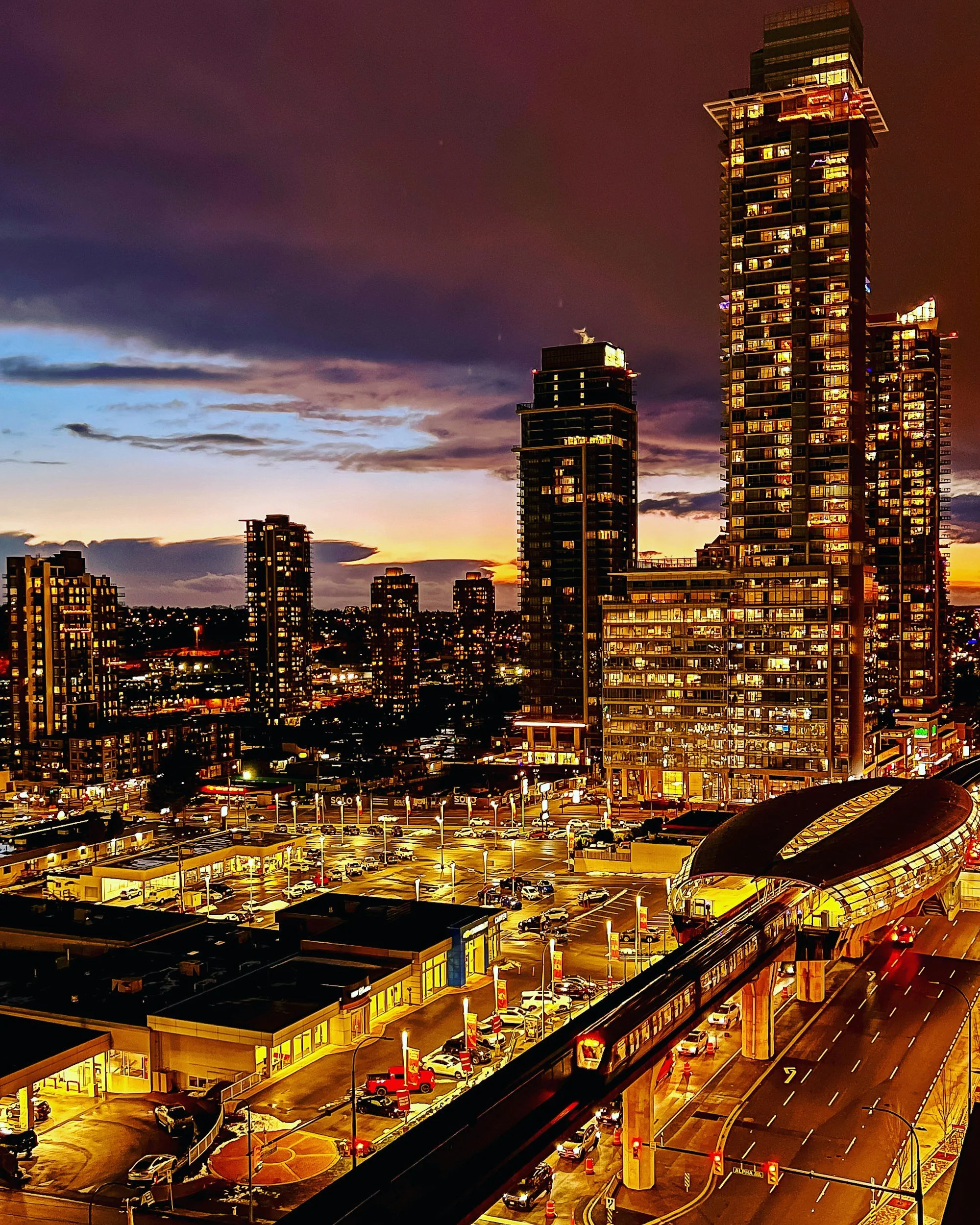 Cityscape at dusk showing tall illuminated skyscrapers, a busy parking lot, and an elevated train track with a train passing through.