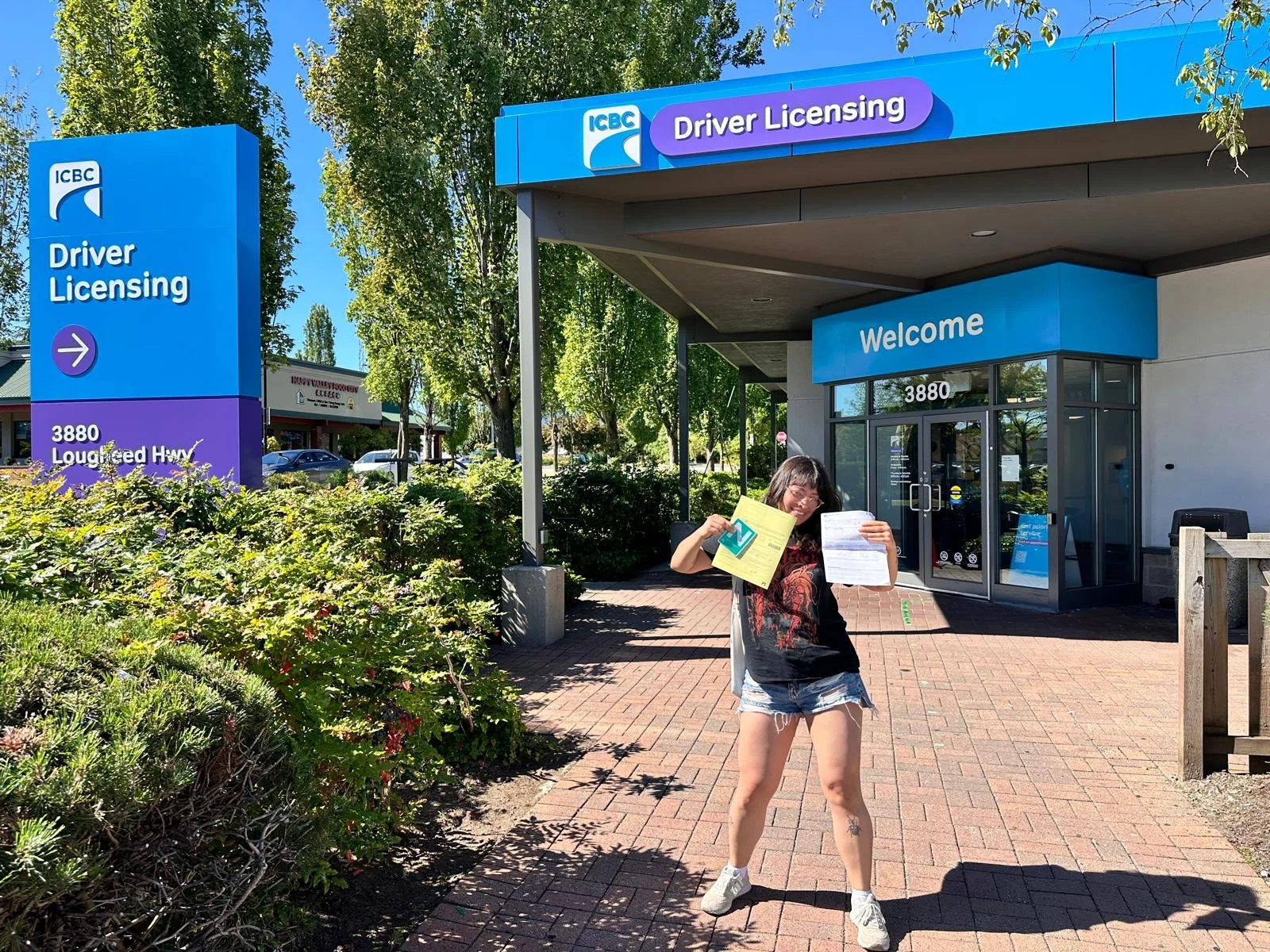 A young woman standing outside the ICBC driver licensing office, holding papers and smiling, with a bright blue sky and green trees in the background.