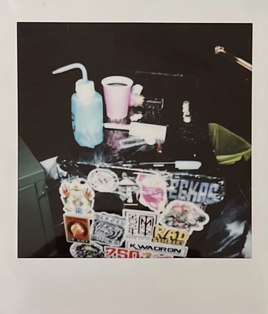 A tattoo station with various stickers, a pink cup, a blue wash bottle, a green container, and miscellaneous items including a tattoo machine and ink bottles.