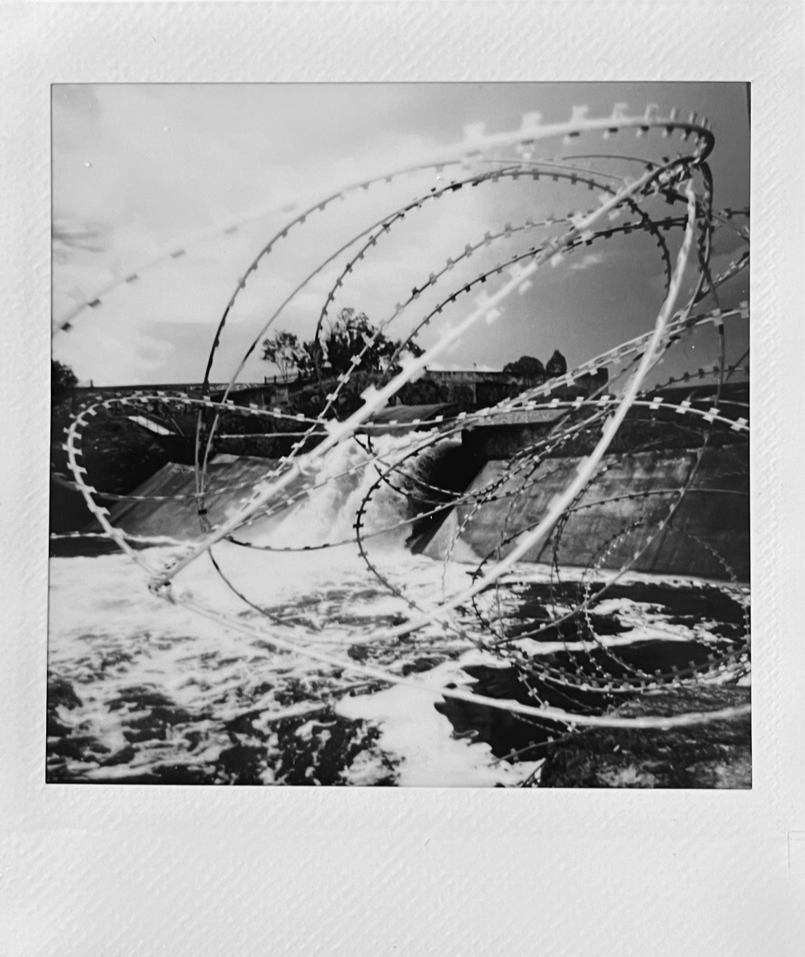 Barbed wire and water spill over a concrete dam or barrier under a cloudy sky.