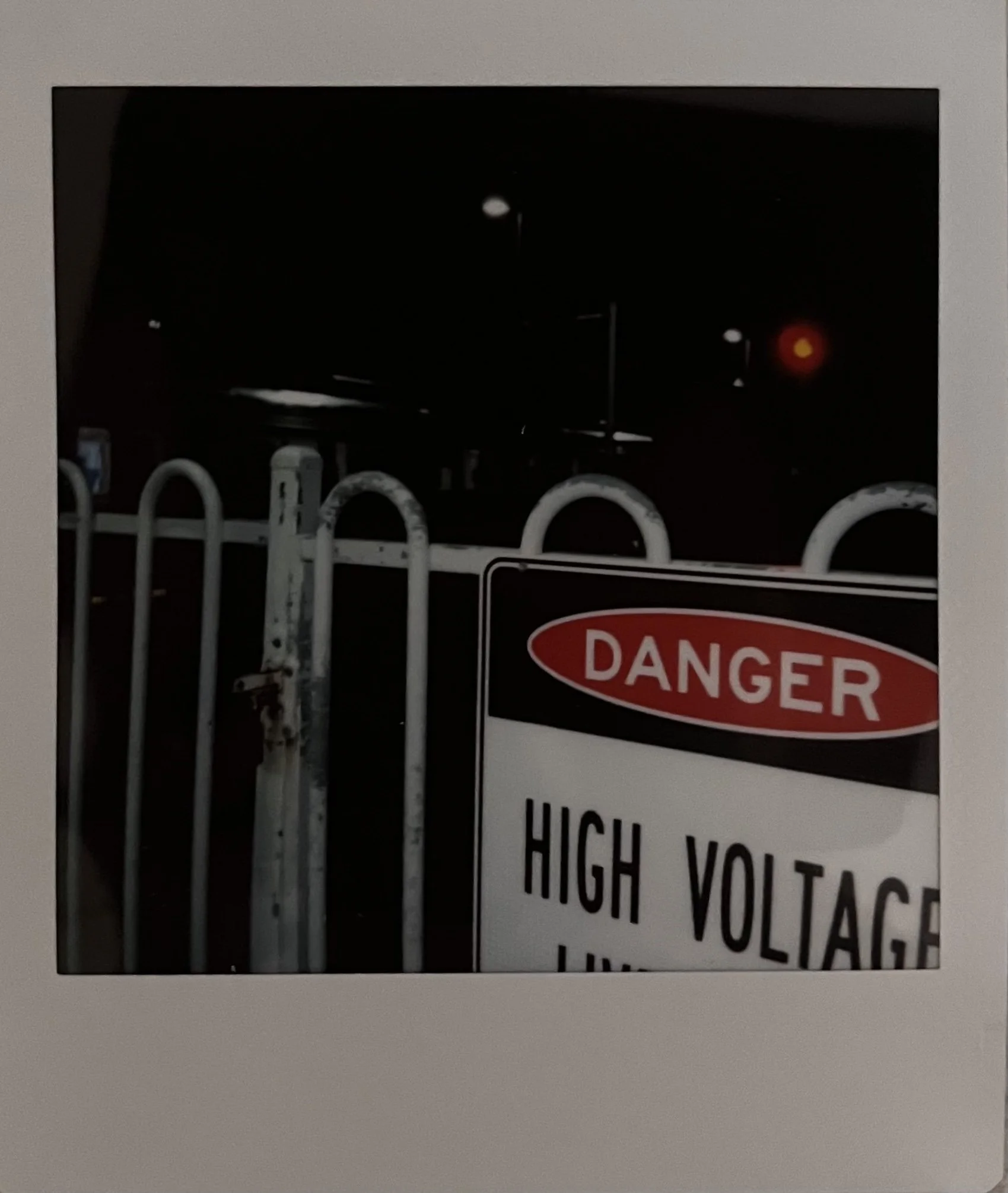 Nighttime scene showing a warning sign that reads "Danger High Voltage" with a metal fence in the foreground and a red traffic light in the background.