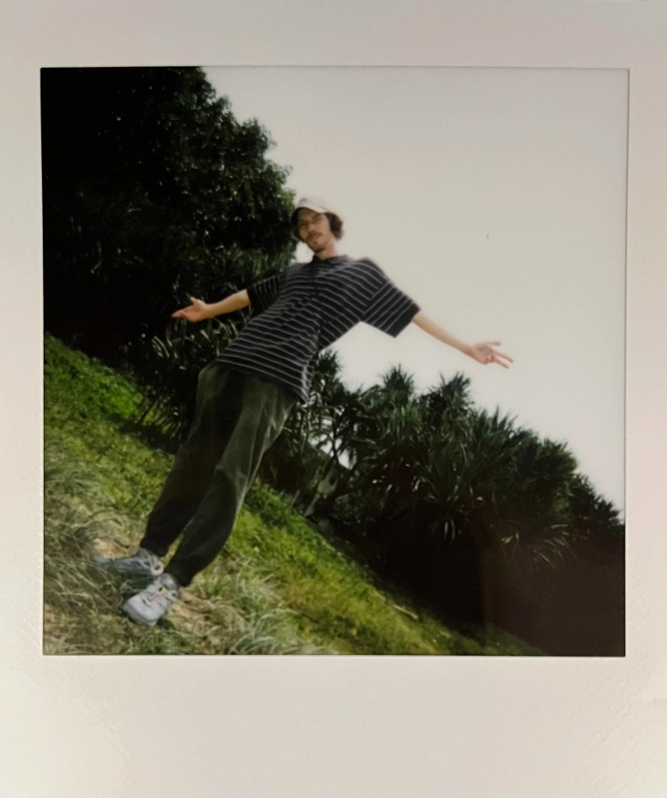 A person standing outdoors on a grassy area with trees in the background, posing with arms outstretched.