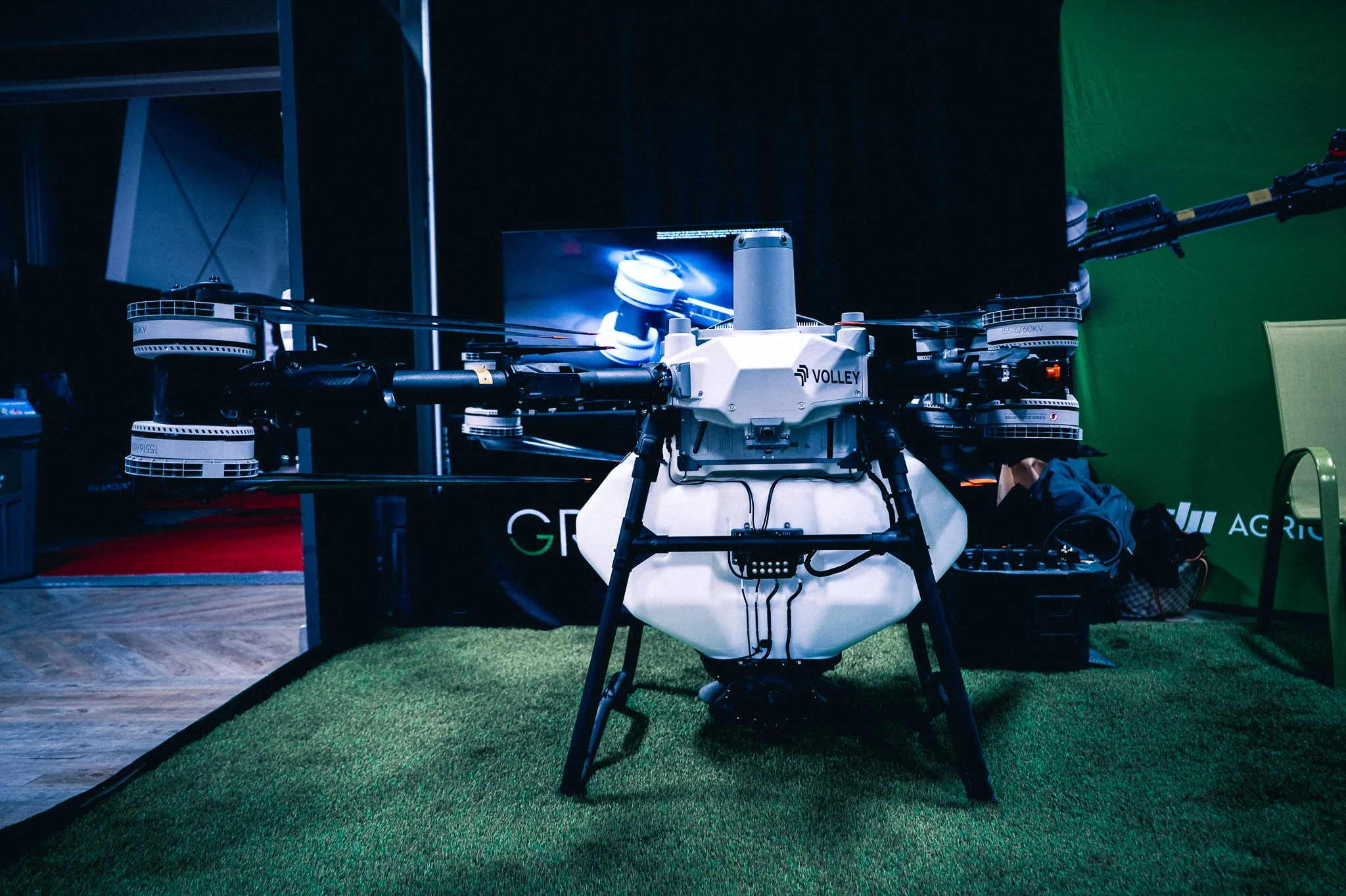 A large drone with six propellers displayed indoors on a patch of artificial grass, with a monitor behind it showing a drone image and a green backdrop on the right side.