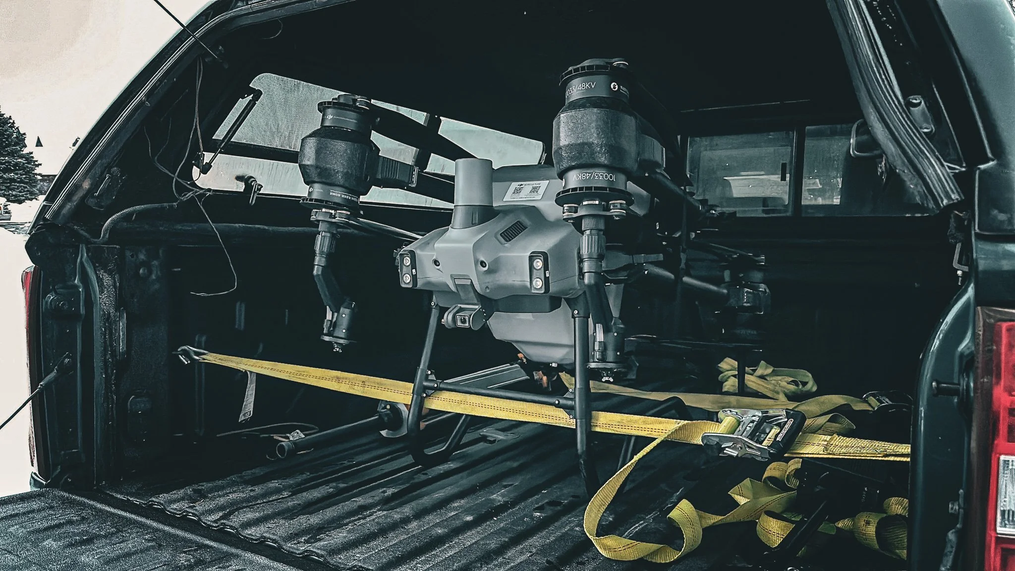 A drone with a gray body and four arms is secured in the back of a black vehicle using yellow straps.