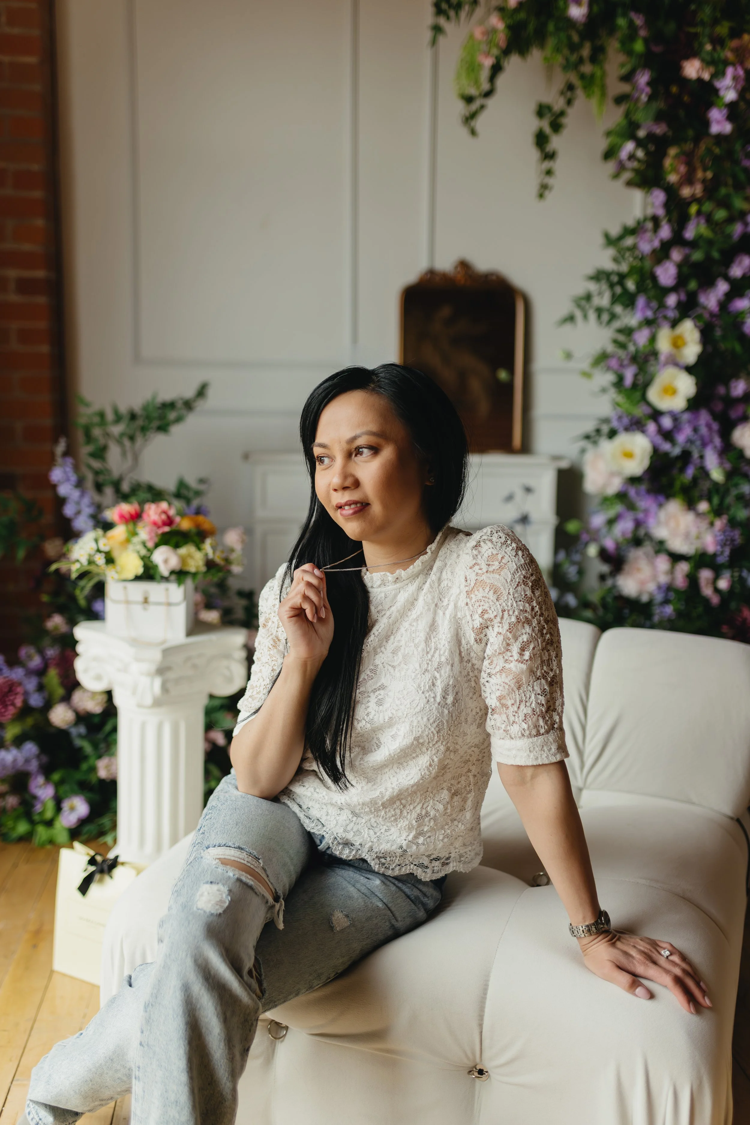 A woman with long black hair sitting on a white sofa surrounded by colorful flowers, wearing ripped jeans and a lace top, holding a necklace, with a thoughtful expression