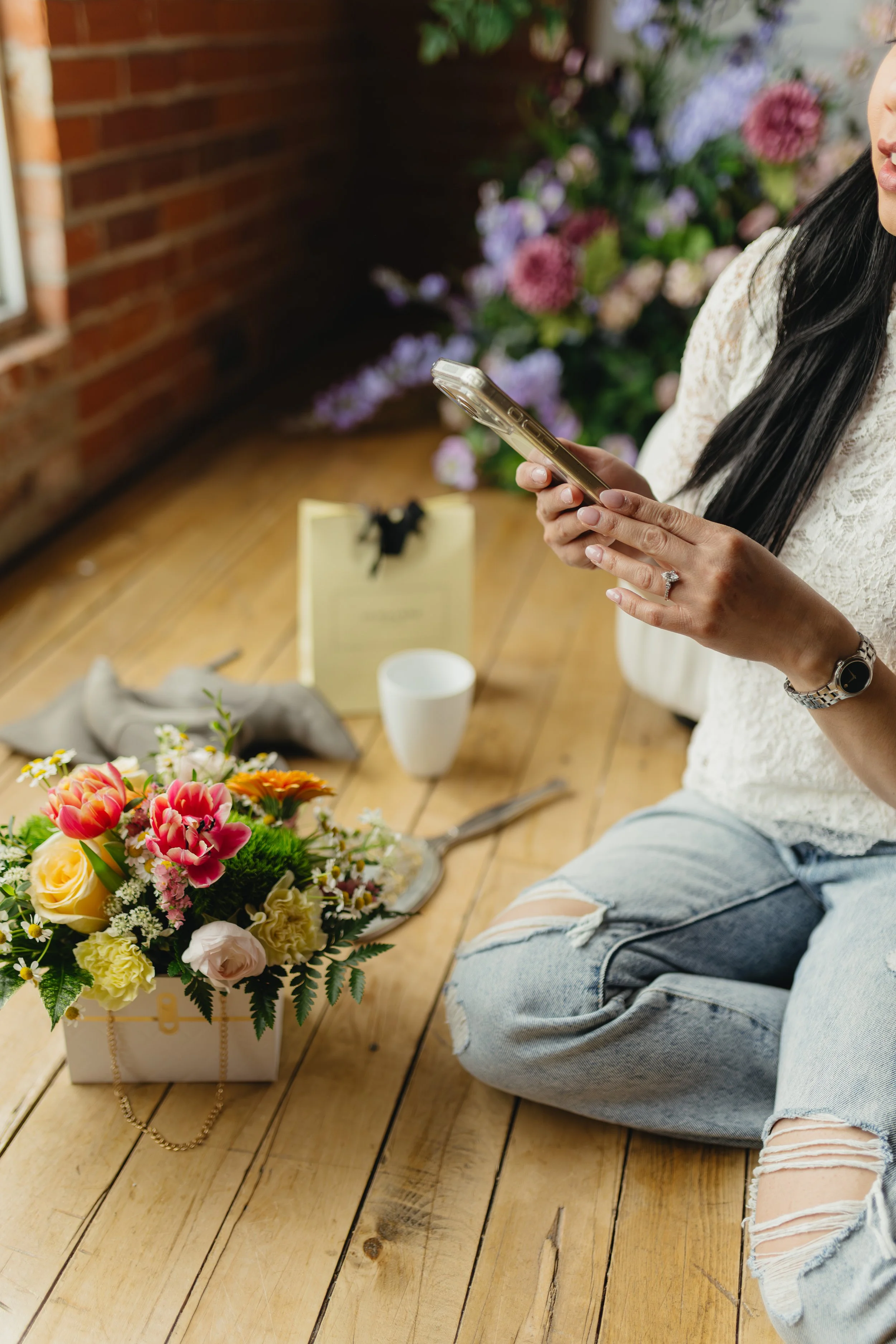 A woman with long black hair wearing a white lace top and ripped jeans, sitting on a wooden floor, holding a smartphone. There is a colorful flower bouquet in a white basket and a yellow gift box with a black bow on the floor in front of her. In the background, there are purple and pink flowers against a brick wall.