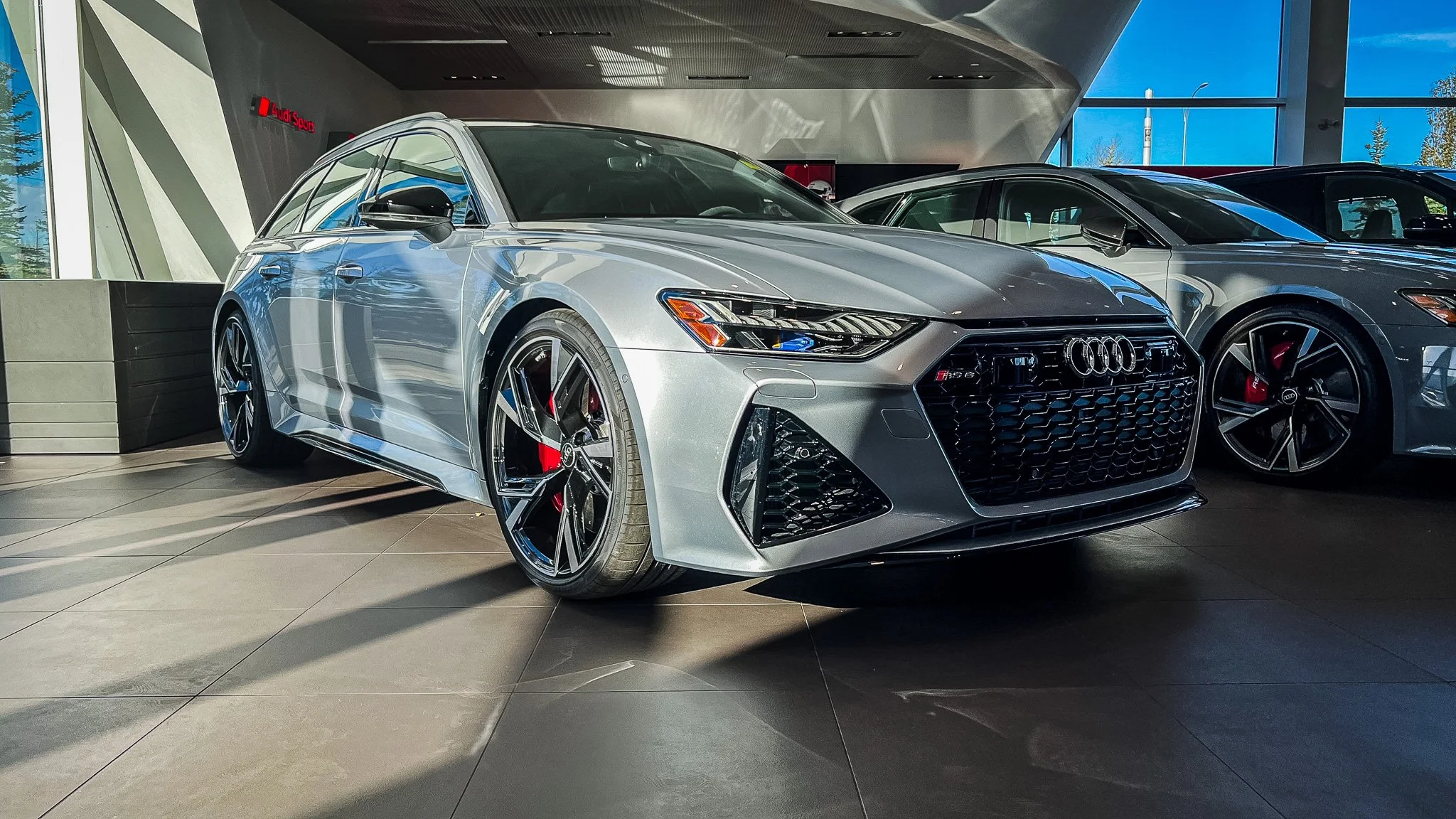 A silver Audi sports sedan on display inside a modern showroom with other vehicles and large windows showing trees outside.