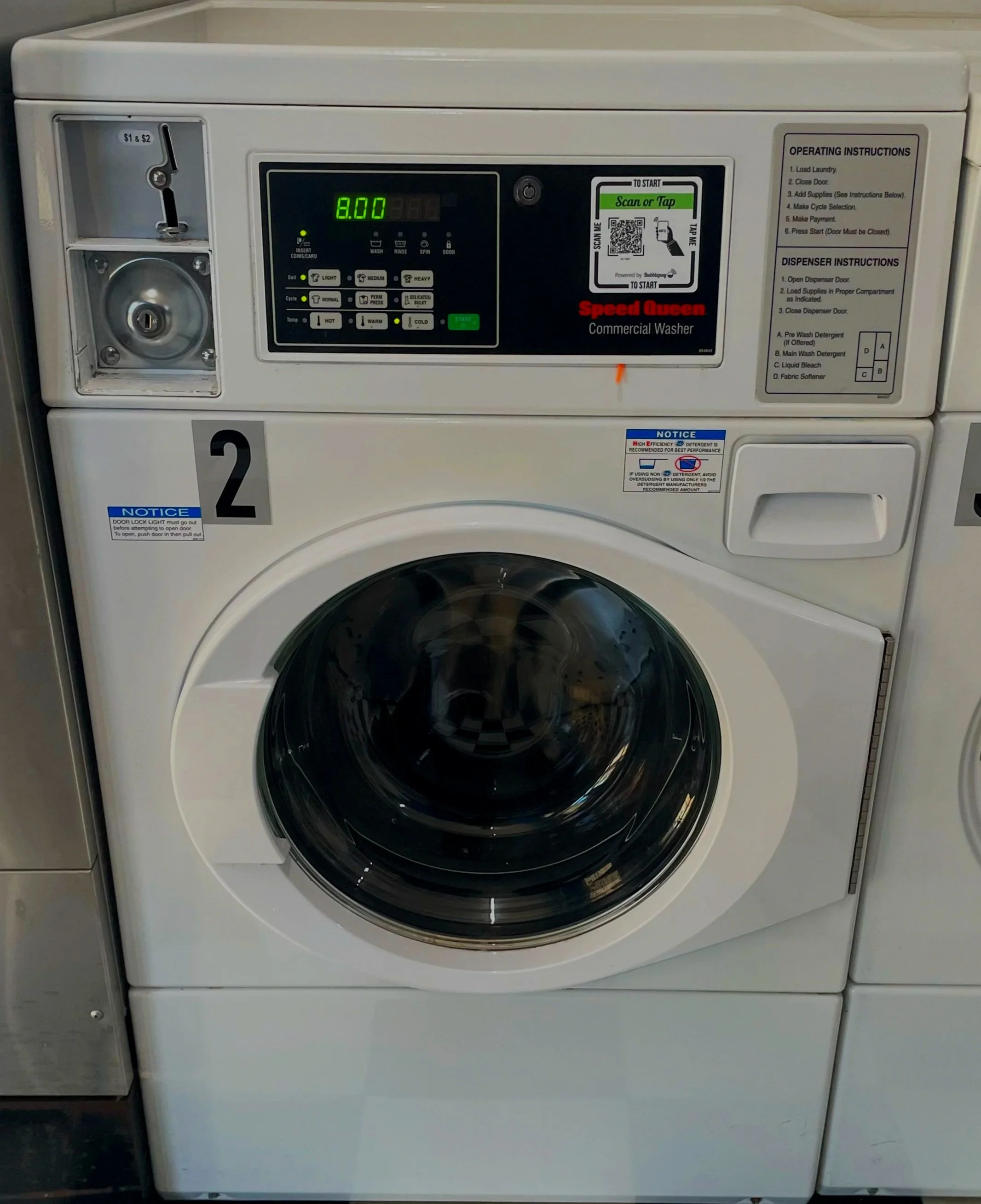 Front view of a Speed Queen commercial washer with digital control panel, coin slot, and instructions, in a laundry facility.