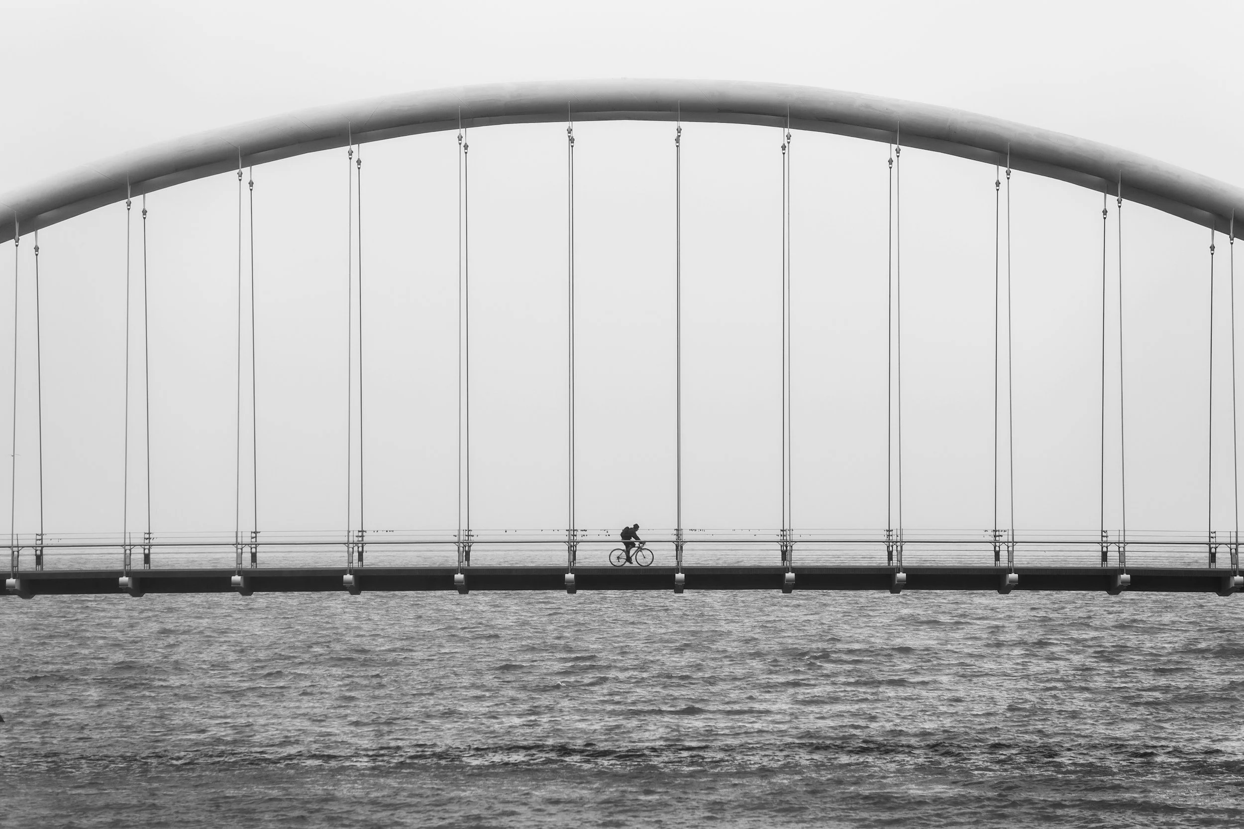 Black and white photo of a person riding a bicycle across a bridge over water, with a large arc structure and suspension cables in the background.