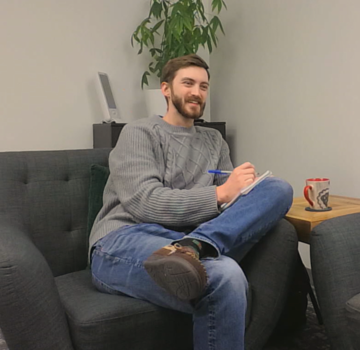 A man with a beard wearing a gray sweater and jeans, sitting on a dark gray couch, holding a pen and notebook, smiling. There is a wooden side table with a patterned mug, a green pillow behind him, and a potted plant with a computer monitor behind. The background has plain light-colored walls.