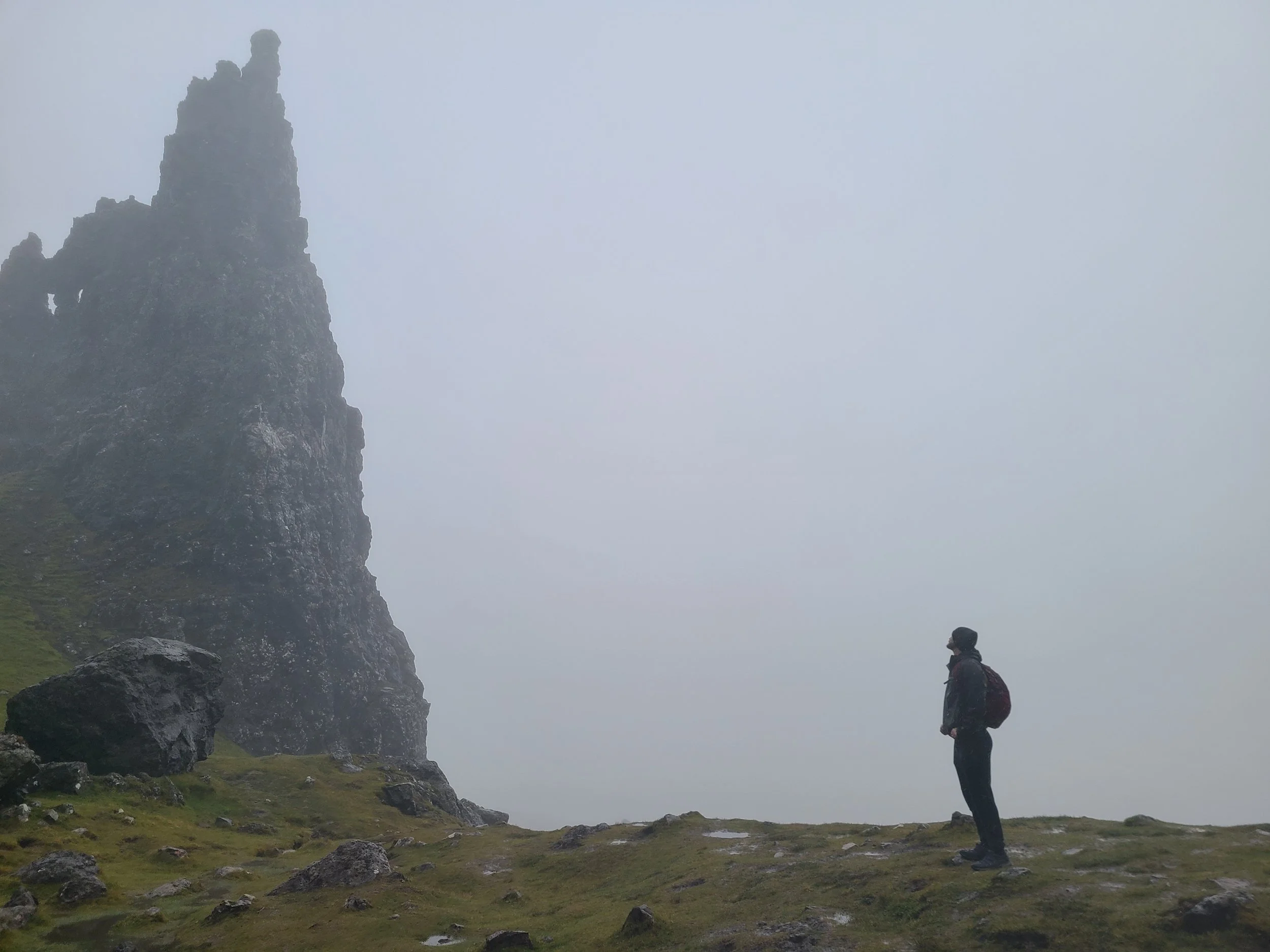A person standing outdoors on grassy terrain with rocks, looking towards a tall, rocky cliff shrouded in mist or fog, during daytime.