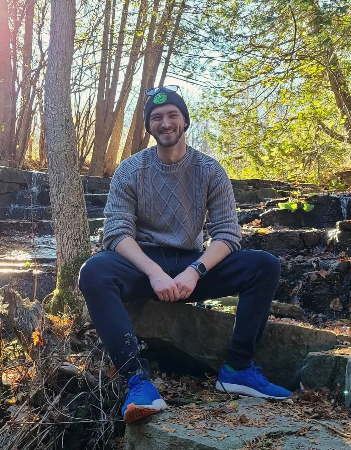 A smiling man sitting cross-legged on a rock in a wooded area, wearing a gray sweater, black pants, blue sneakers, a black beanie, and sunglasses on his head, with a stream and trees in the background.