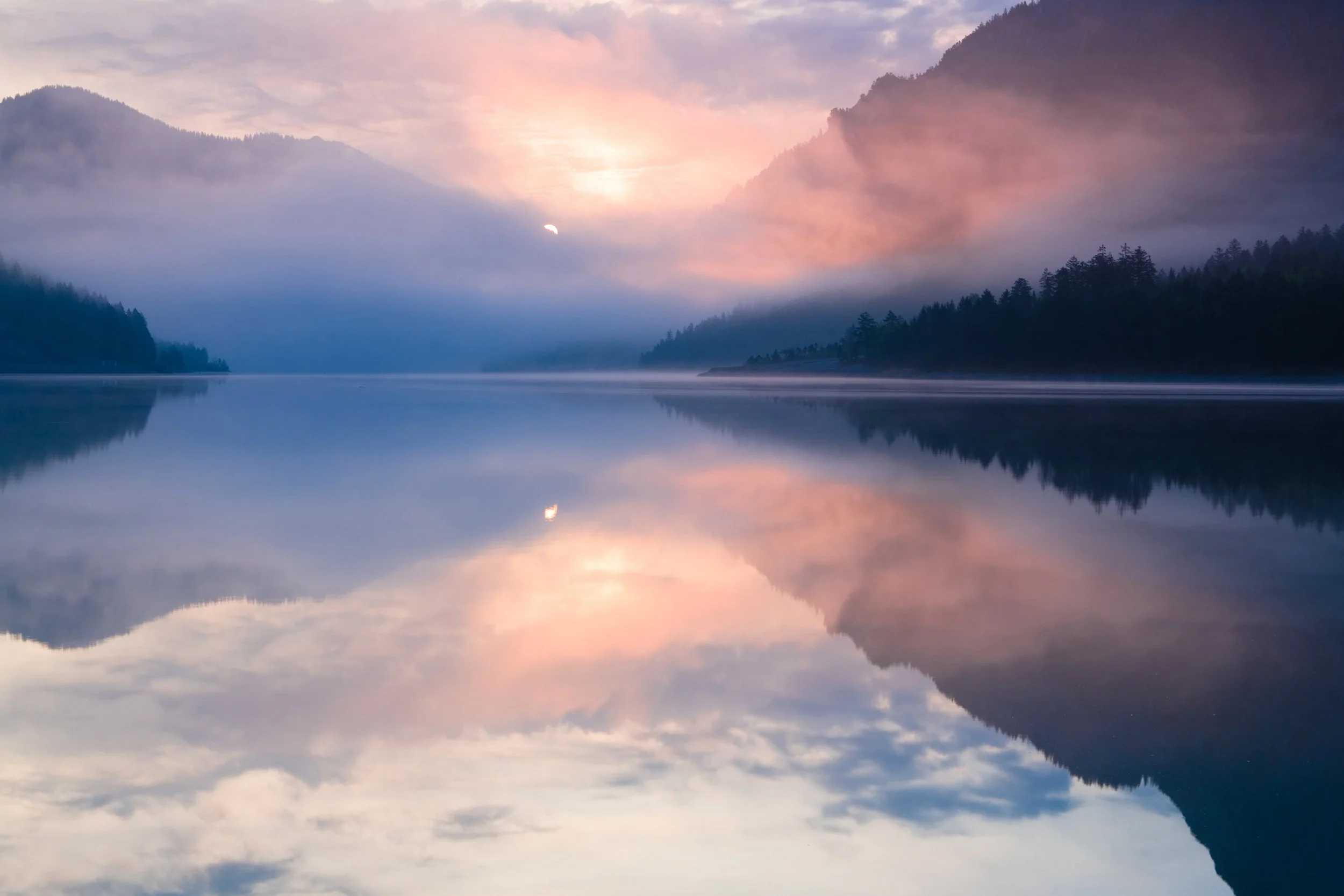 Sunrise over a misty lake with reflection of mountains and sky in the water.