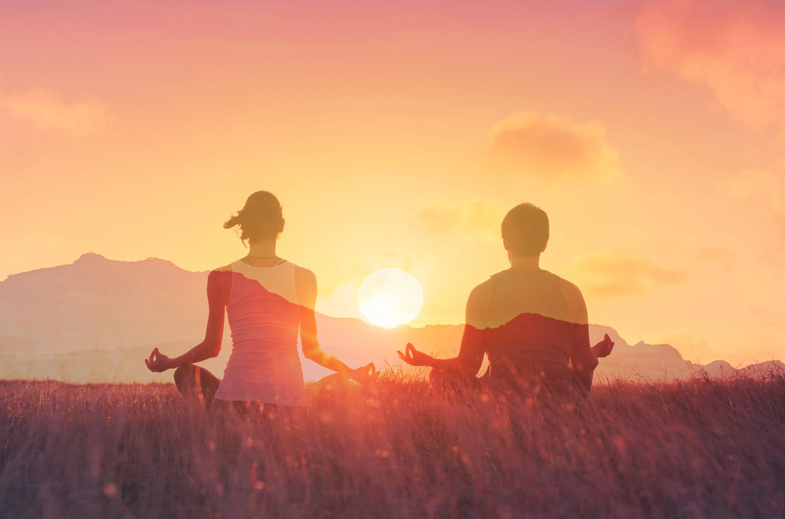 Two people practicing meditation or yoga in a field during sunset, with mountains in the background.