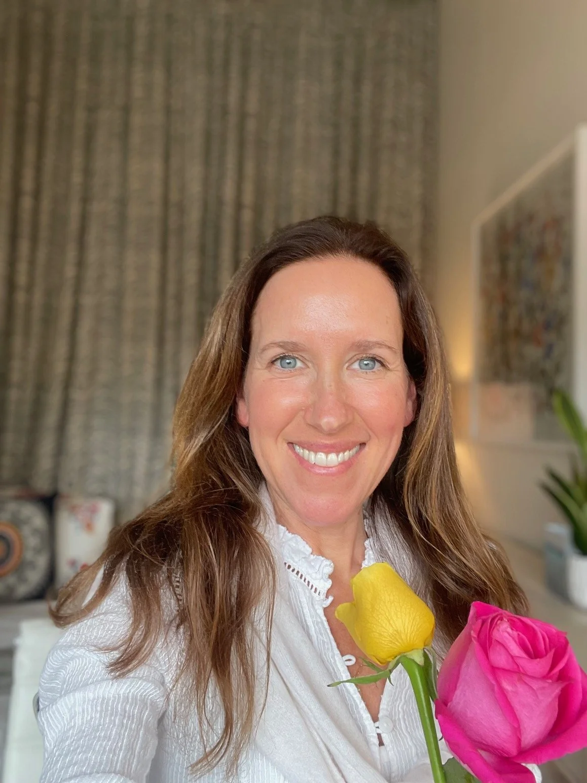 A woman with long brown hair and blue eyes smiling, holding a yellow tulip and a pink rose in front of a blurred indoor background.