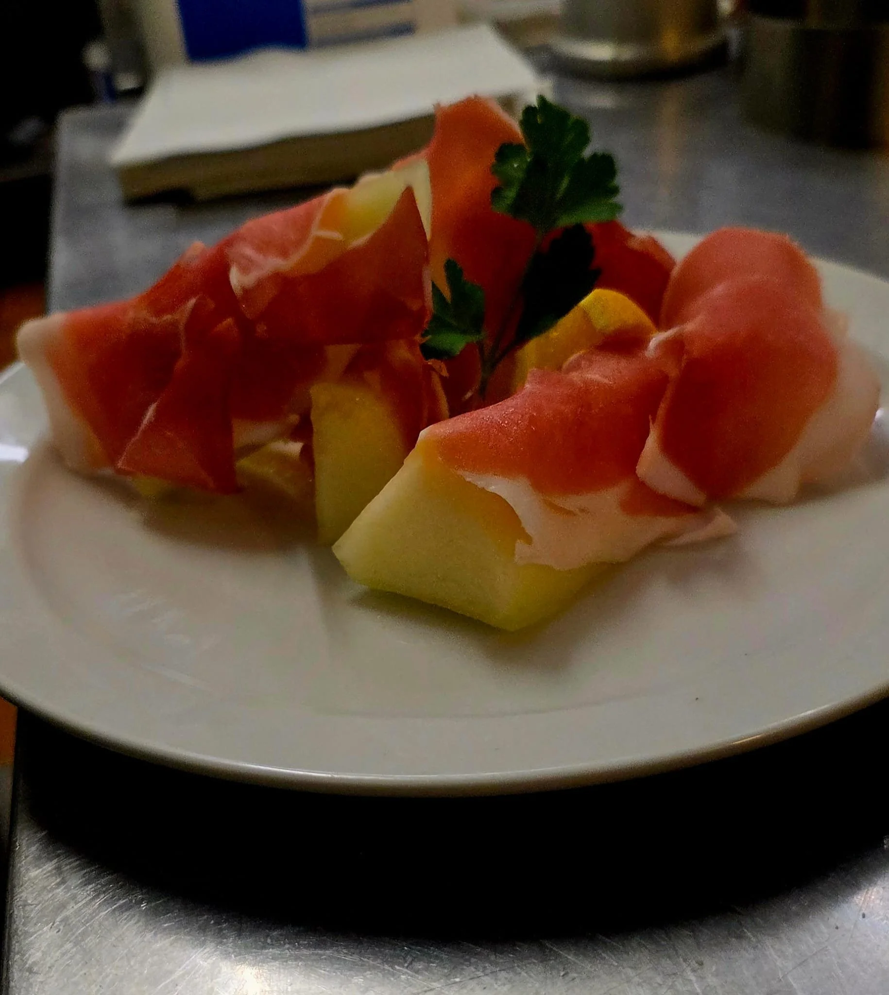 A white plate holding pieces of cantaloupe melon wrapped in prosciutto and garnished with a sprig of parsley.