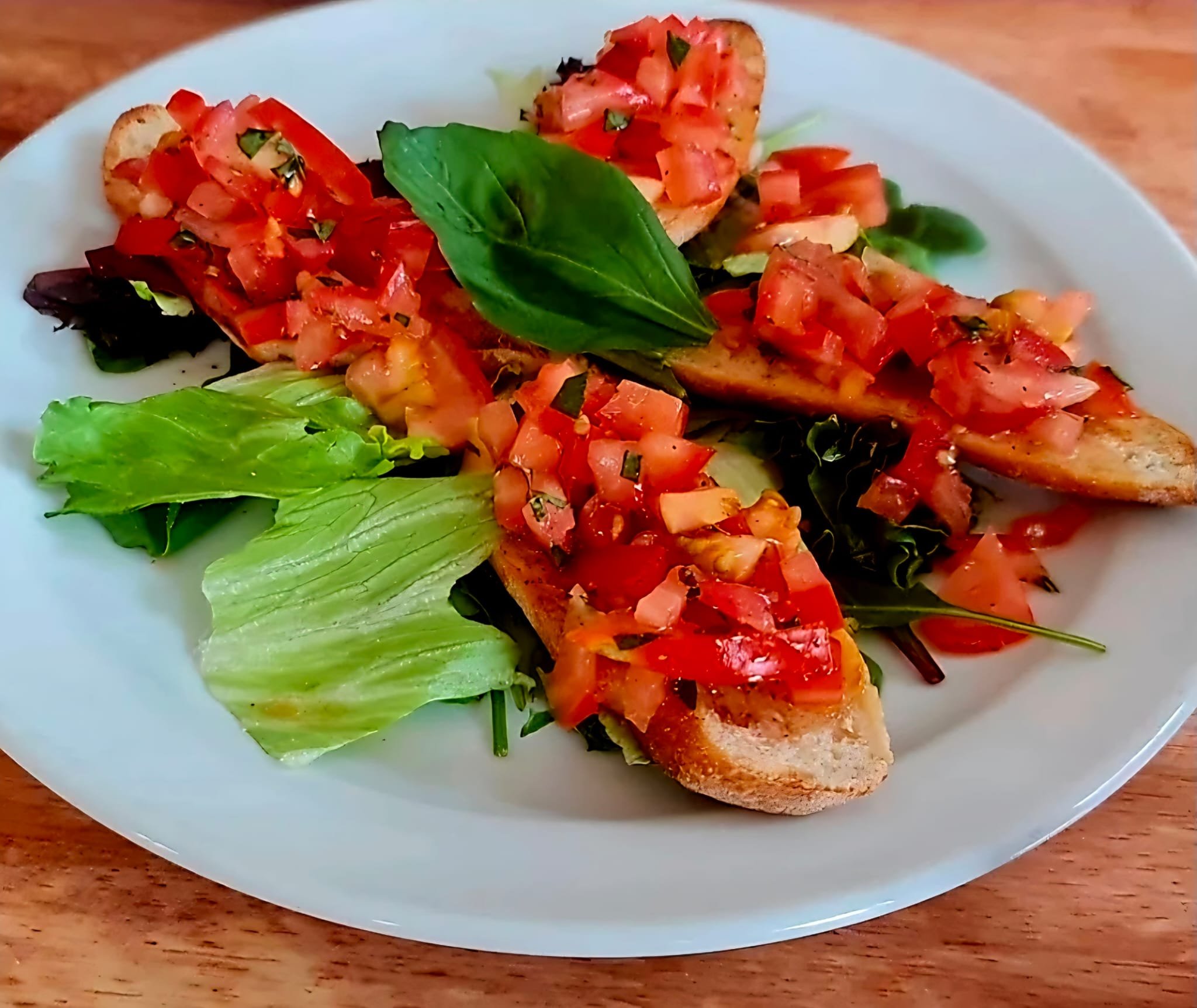 Plate of bruschetta topped with chopped tomatoes, basil, and olive oil, served on toasted Italian bread with leafy greens on the side.