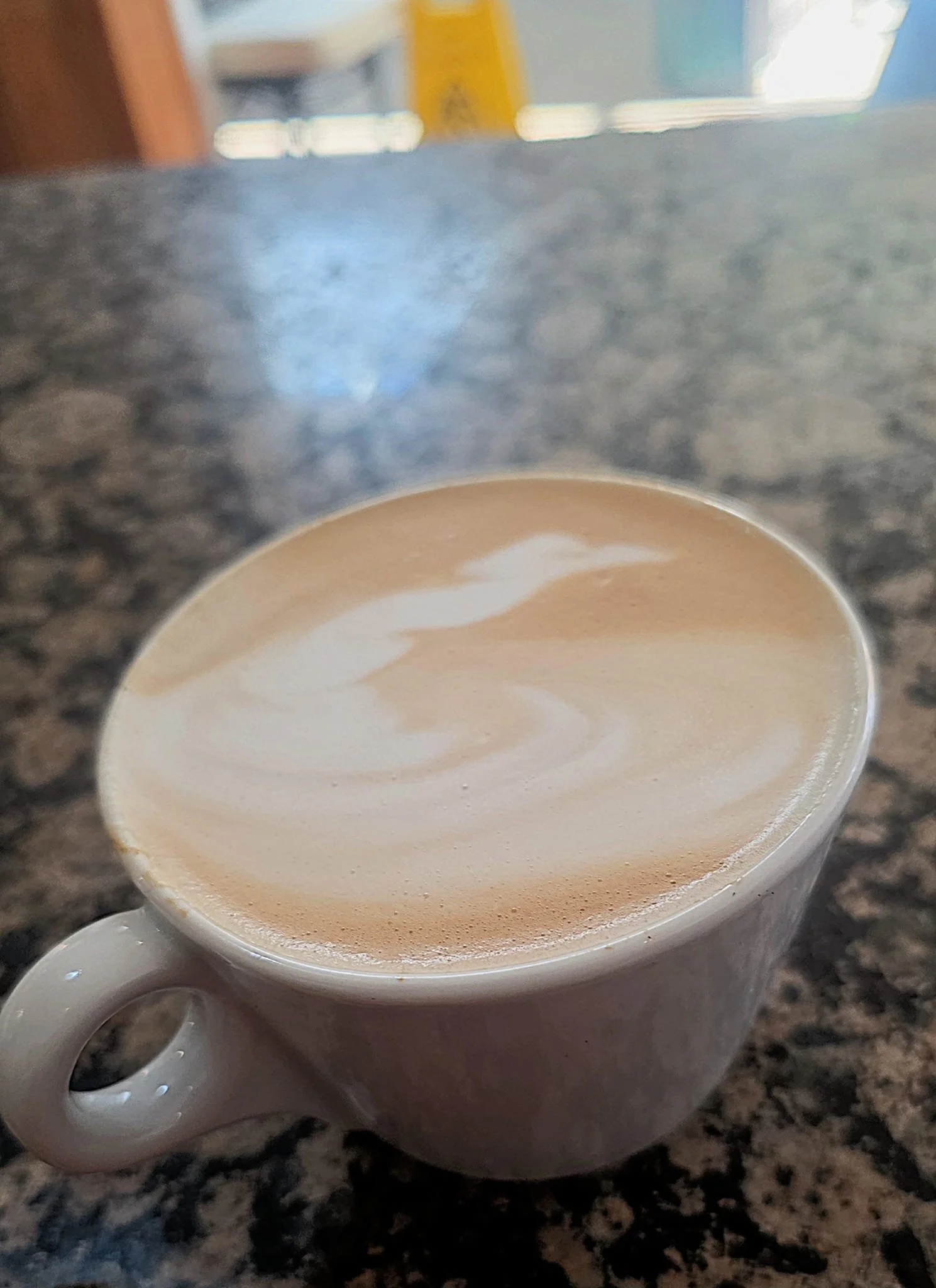 A white ceramic mug filled with a layered latte, with steamed milk and foam art on top, placed on a dark granite countertop.