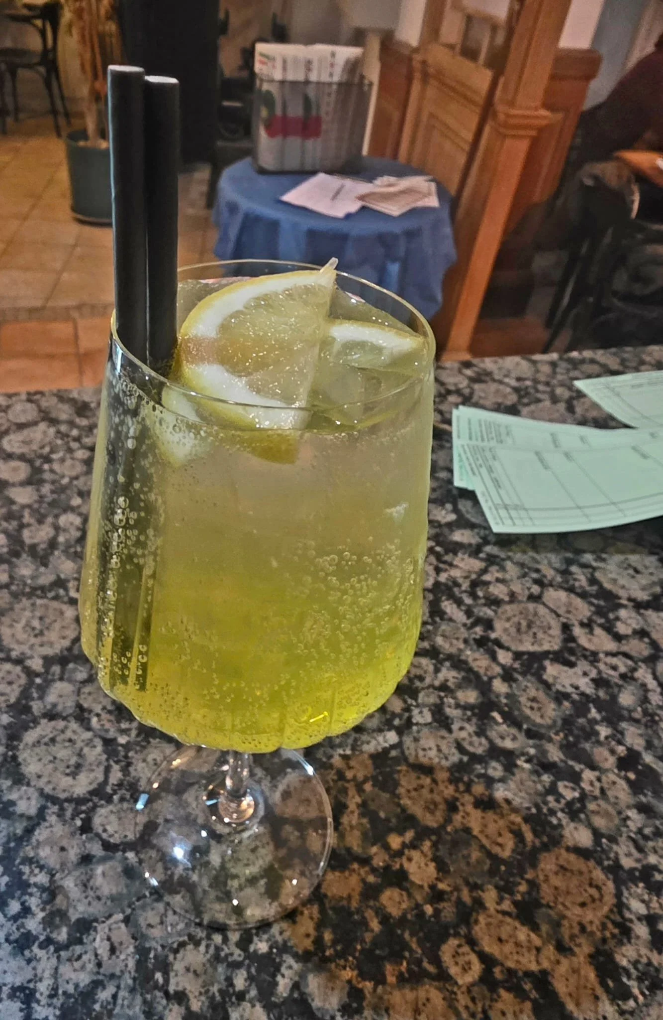 A glass of lemon soda with ice and two black straws on a granite bar counter in a restaurant.