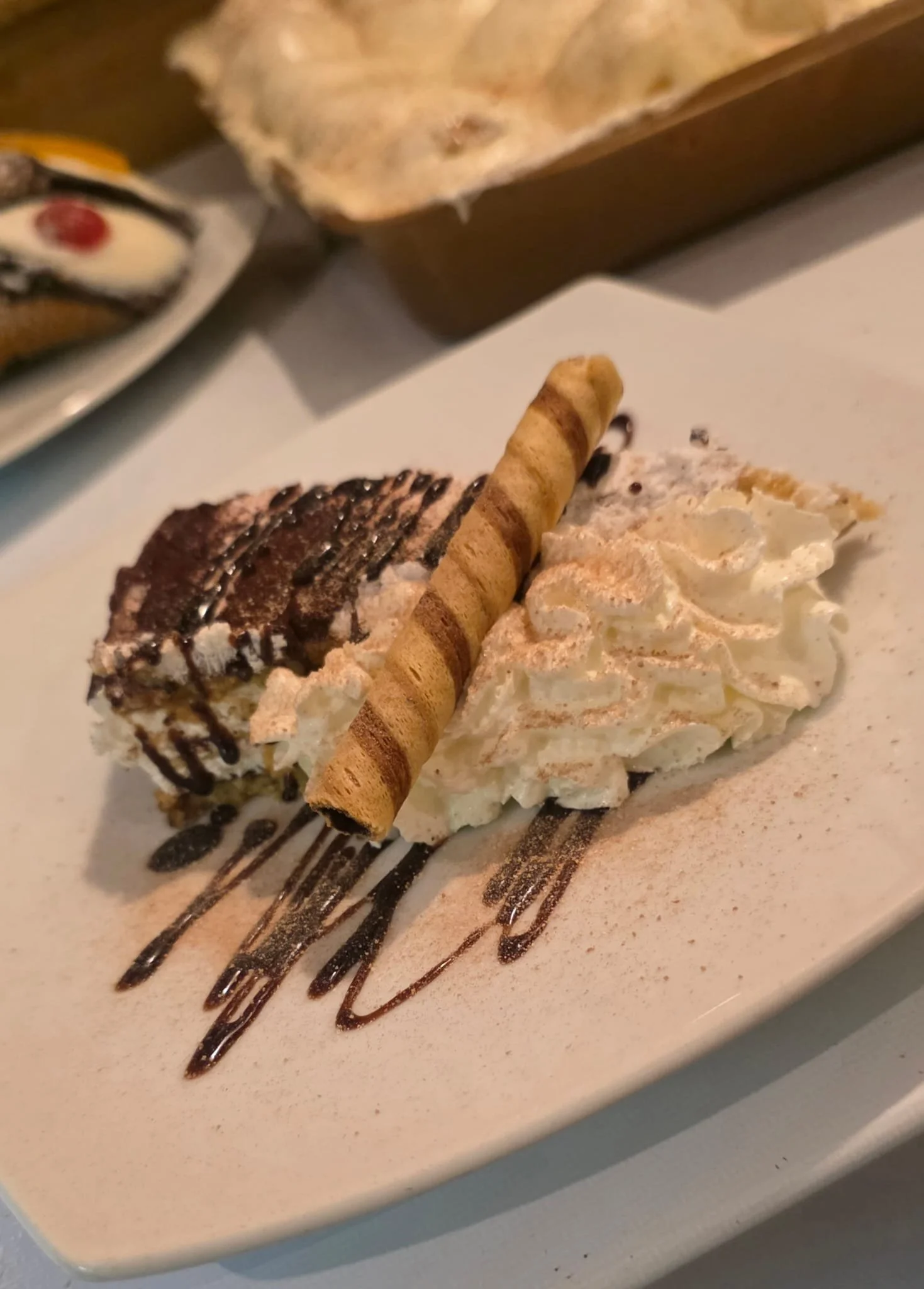 Slice of chocolate cake with whipped cream, drizzled with chocolate syrup, garnished with a rolled wafer cookie, served on a white plate with additional whipped cream and chocolate drizzle.