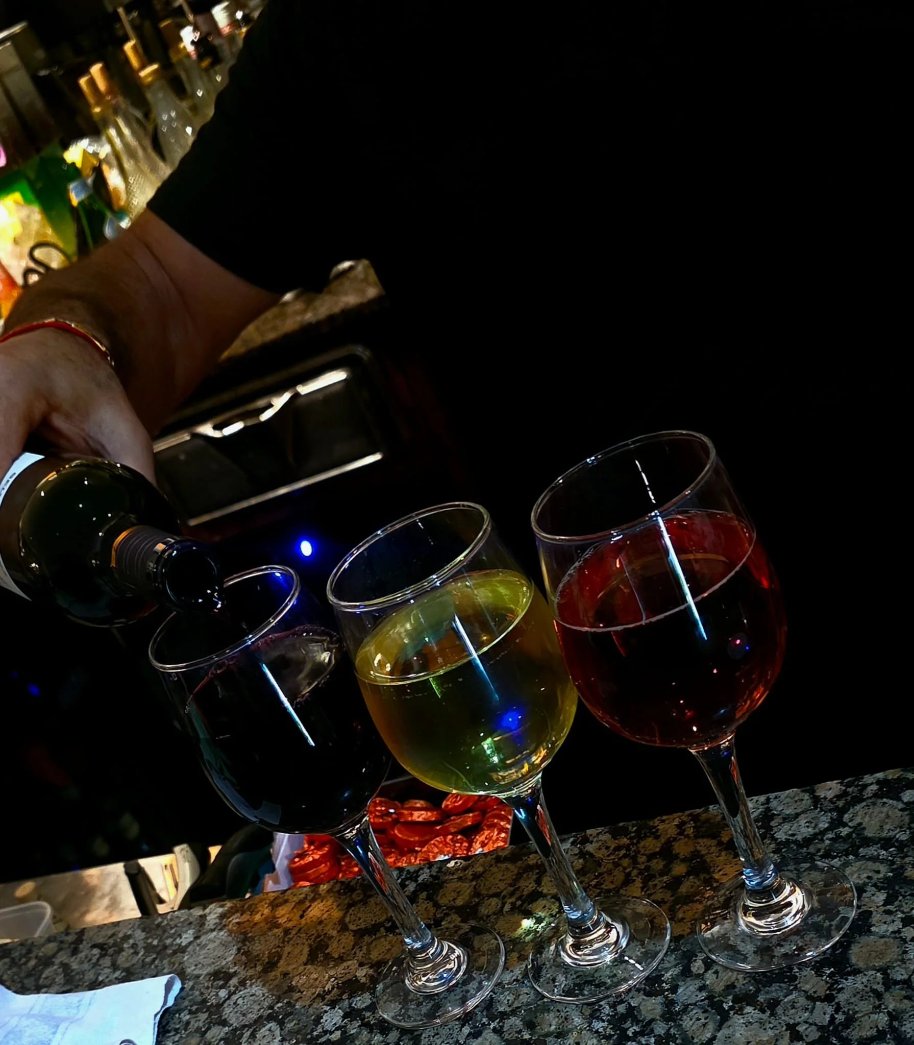 Three wine glasses filled with different colored wines (red, white, and dark red) on a bar counter, with a person pouring a drink in the background.