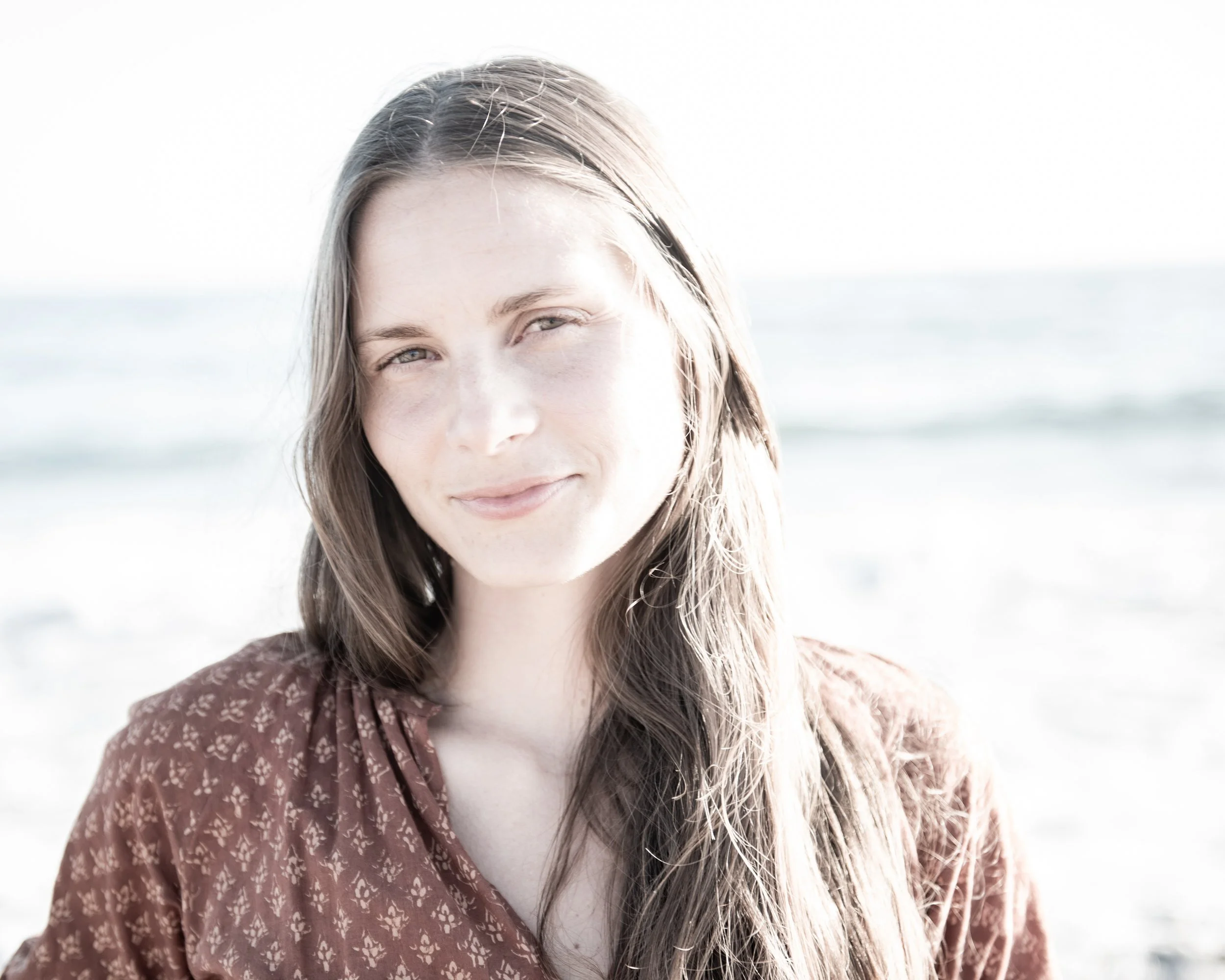 Close-up of a young woman with long light brown hair, smiling softly, outdoors near the water on a bright day.