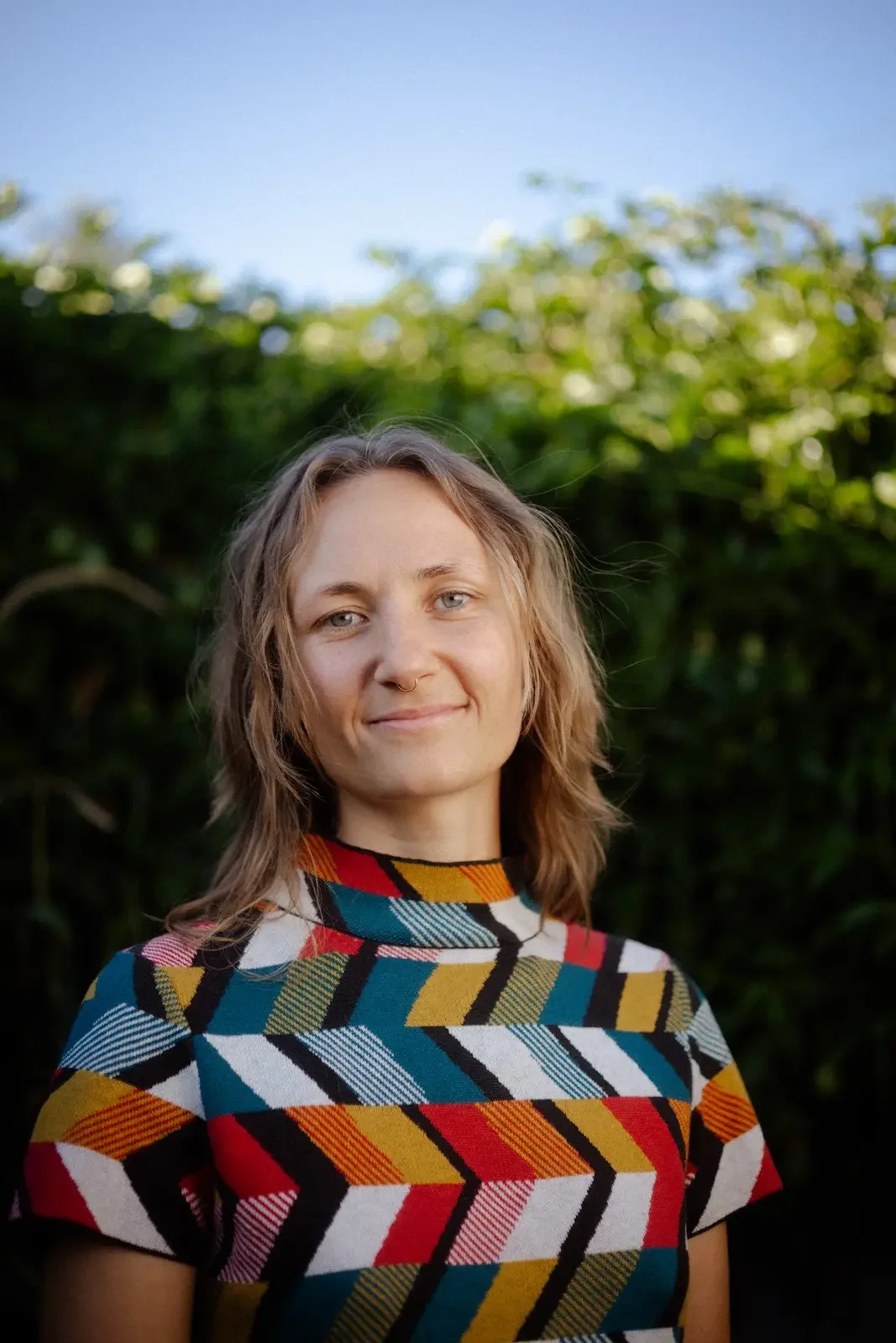 A woman with blonde hair and light-colored eyes standing outdoors in front of greenery and a blue sky, wearing a colorful geometric patterned shirt.