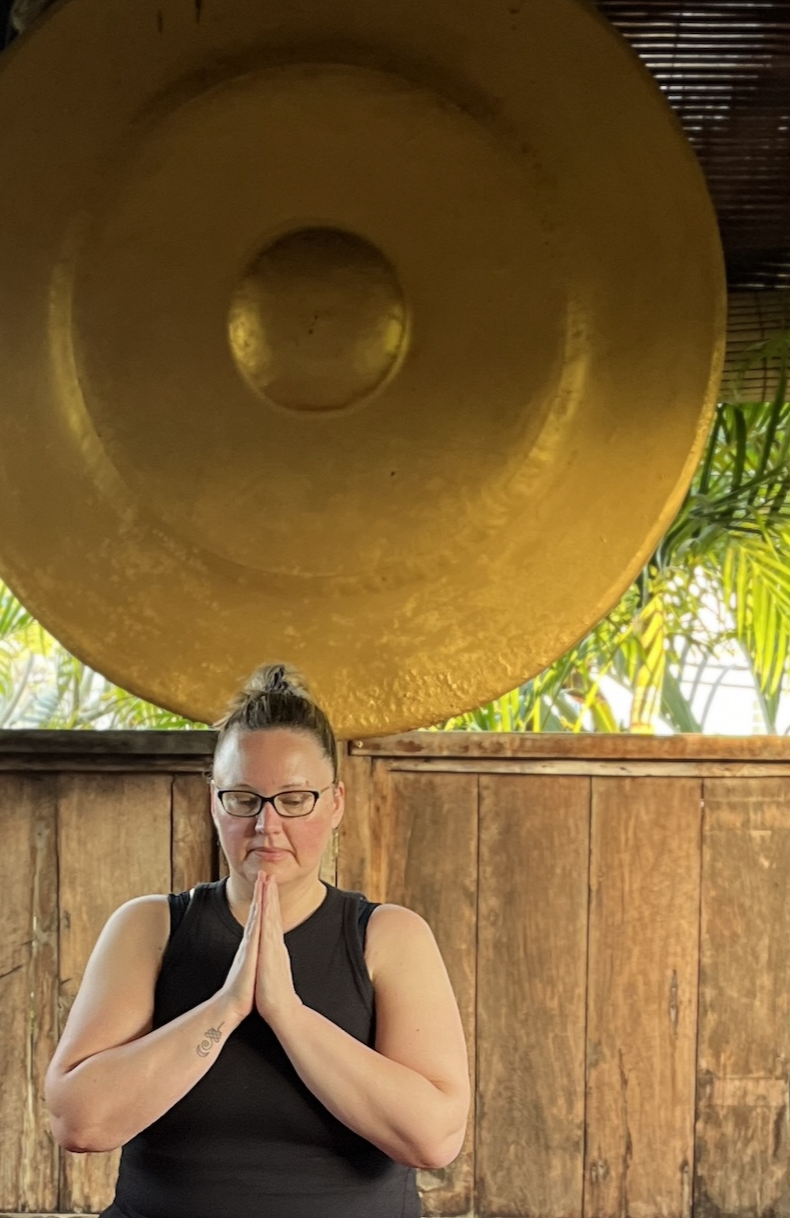 A woman practicing yoga or meditation with her hands in prayer position, standing in front of a large gong, surrounded by tropical plants.