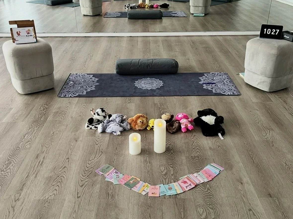 A calm children’s yoga studio with a yoga mat, bolster, soft floor seating, plush toys, LED candles, and mindfulness cards arranged in a welcoming circle.
